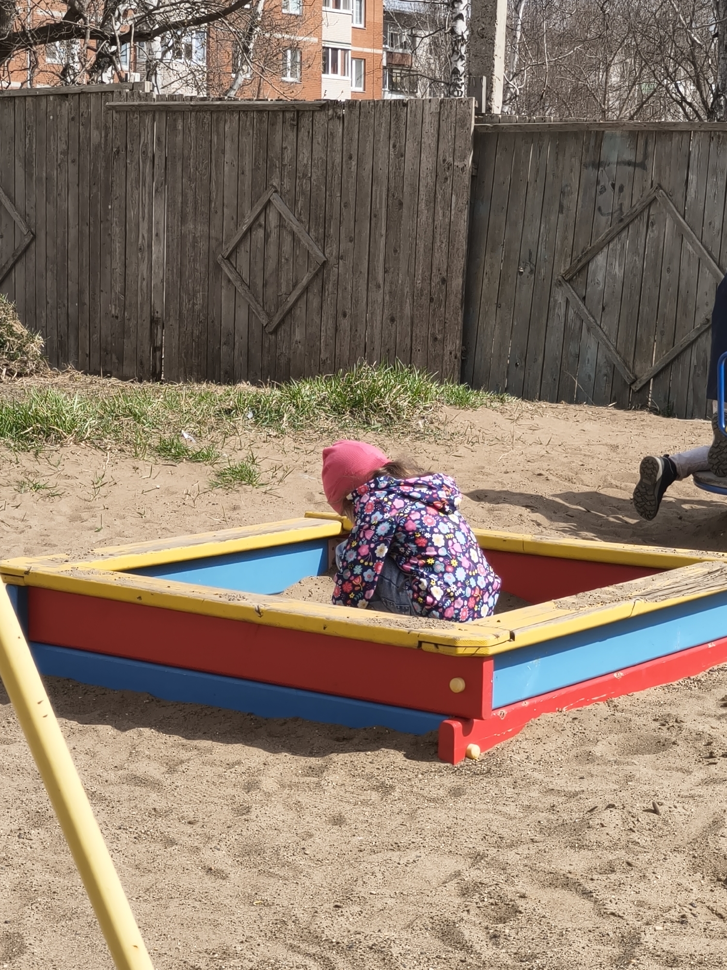 A child in a pink hat and a floral jacket is in a colorful square sandbox filled with sand. Another person, likely an adult, is visible in the background, slightly out of focus. Behind the sandbox is a weathered wooden fence with diamond-shaped decorations. In the distance, there is a multi-story building with windows, partially obscured by bare tree branches. The scene appears to be set in a playground or a park.