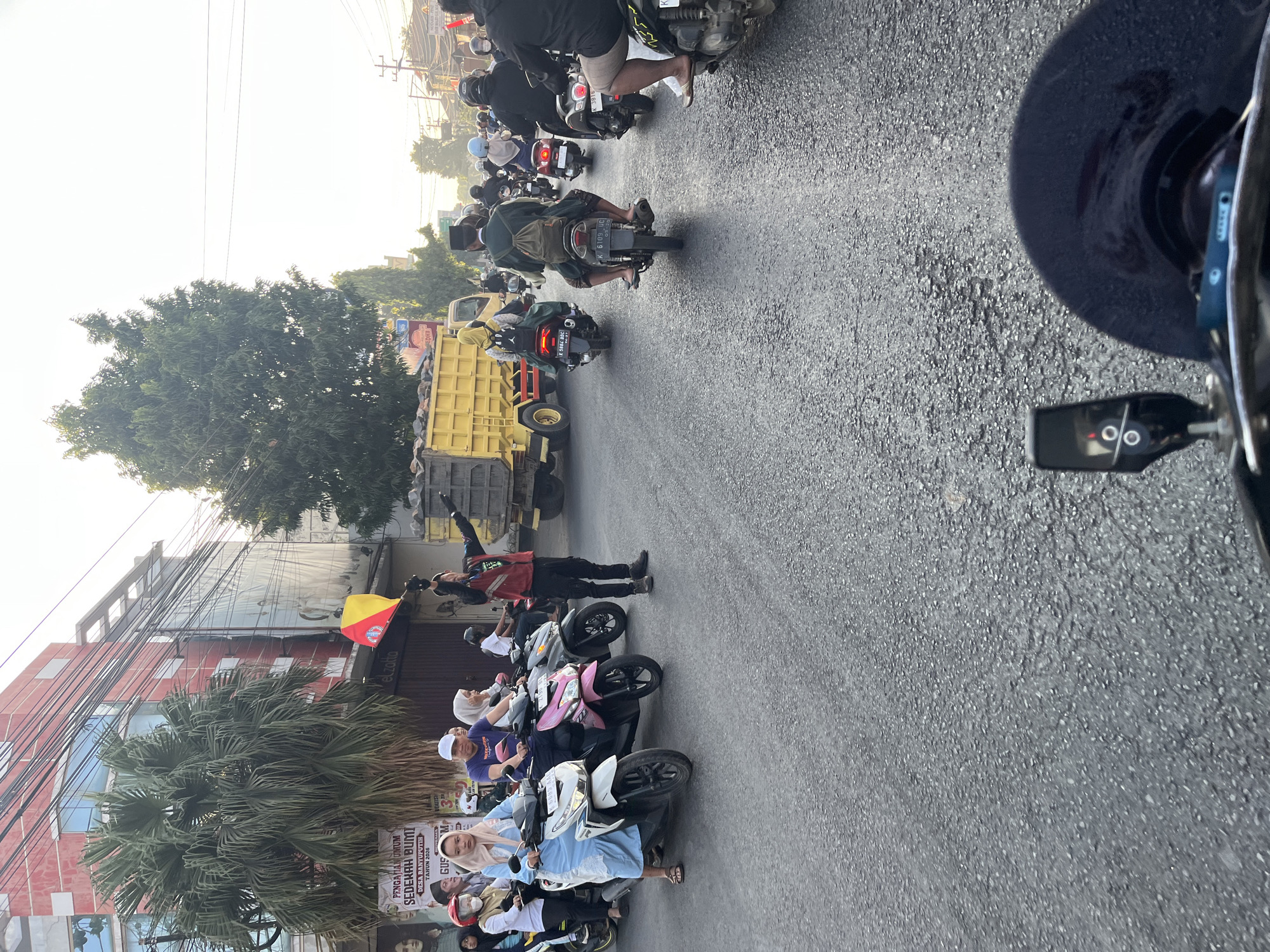 The image depicts a street scene in Banyuputih, Indonesia, with a convoy of motorcycles. A person in a red vest and black pants is standing in the middle of the road, holding a yellow and red flag, appearing to direct traffic. Several motorcycles with riders are present, some carrying passengers. On the left side of the road, a building with a red facade and large windows stands, with a palm tree in front of it. A yellow truck is visible further down the road, also part of the convoy. The road surface is asphalt. The sky is bright, suggesting daytime.