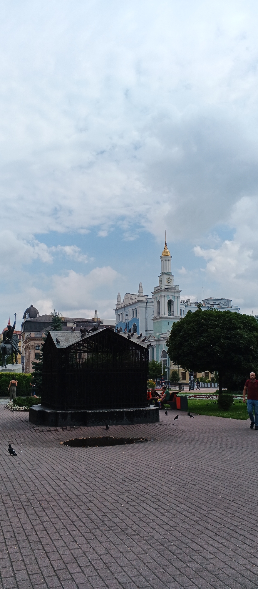 The image depicts an outdoor public space in Kyiv, Ukraine, likely a square or park. In the foreground, a dark, cage-like structure is situated on a paved area made of red brickwork. Several pigeons are perched on top of and around the structure. To the left, a statue of a person on horseback is visible, with buildings in the background. Dominating the background is a light blue and white building with a tall clock tower topped by a golden spire. A large, dark green tree stands to the right of the cage-like structure. A few people are scattered throughout the scene; one man in a red shirt and blue jeans walks away from the viewer on the right side of the frame, and a group of people are seated on a bench on the left side of the frame. The sky is overcast with white and gray clouds.