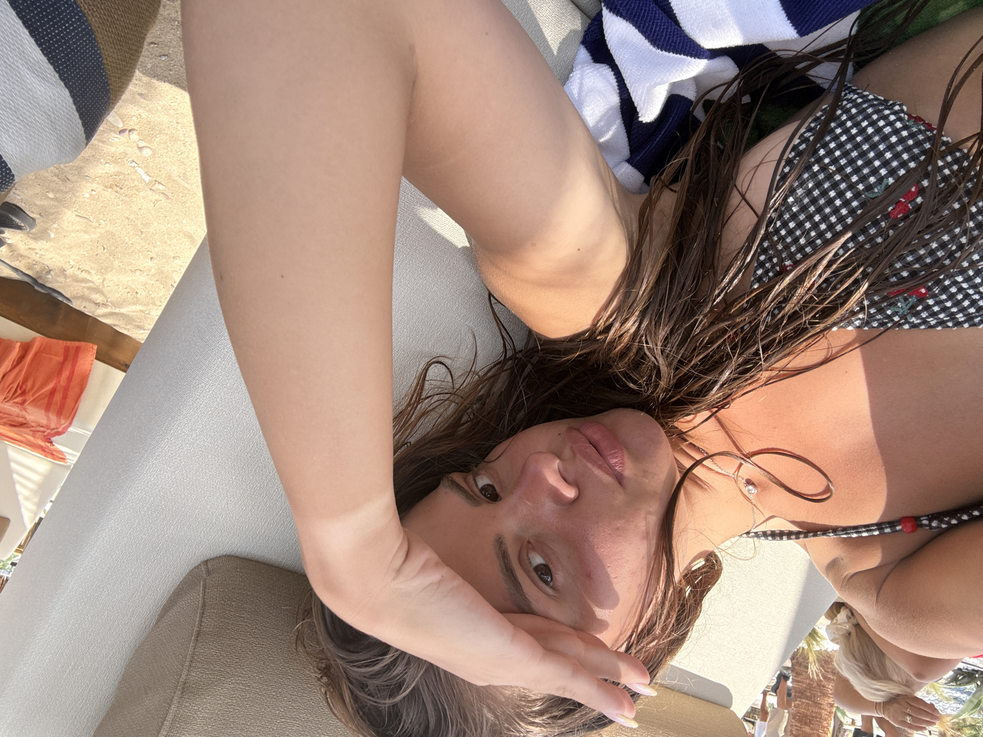 A young woman with long, wet brown hair is lying on a lounge chair outdoors, with her hand shielding her eyes from the sun. She is wearing a black and white checkered bikini top with red embroidered cherries and a silver necklace with a small diamond pendant. A striped towel is draped across her lower body. The background shows a sandy beach area with a few small pebbles, and in the distance, blurred figures of people and palm trees suggest a beach resort or promenade. The overall setting appears to be Ofrynio Beach, Greece, during daylight hours.
