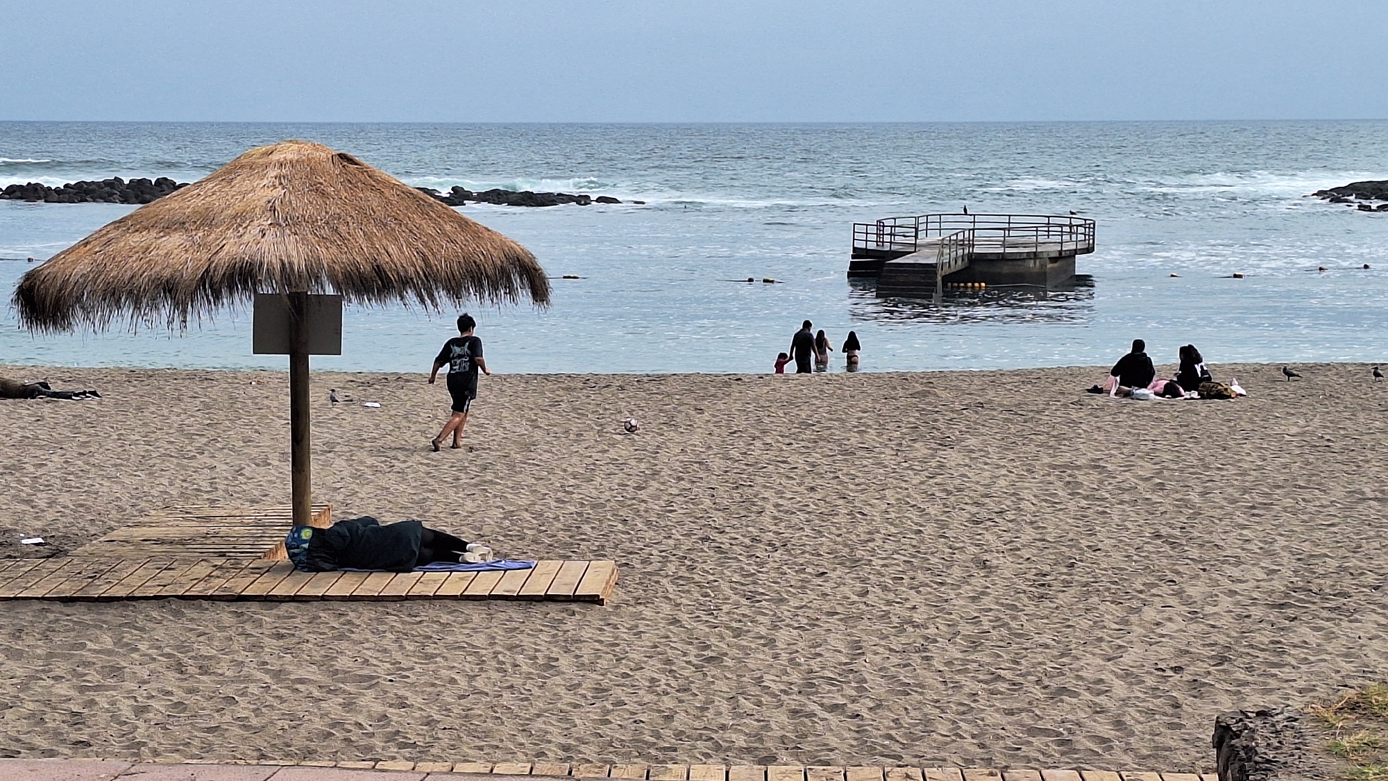 The image depicts a beach scene on an overcast day in Arica, Chile. In the foreground, a thatched umbrella stands over a wooden platform where a person is lying down, covered by a dark blanket, with their head on a blue towel. To the right of the umbrella, a person in a black t-shirt and shorts is running on the sand. Further back, a soccer ball rests on the sand. In the water, a family of four, including two adults and two children, are wading. A wooden pier extends into the sea with a platform at the end. On the far right of the beach, two individuals are sitting on a blanket, appearing to relax. Several birds are present on the sand. The ocean, with moderate waves, stretches to the horizon under a gray sky. Rocky outcrops are visible in the water.
