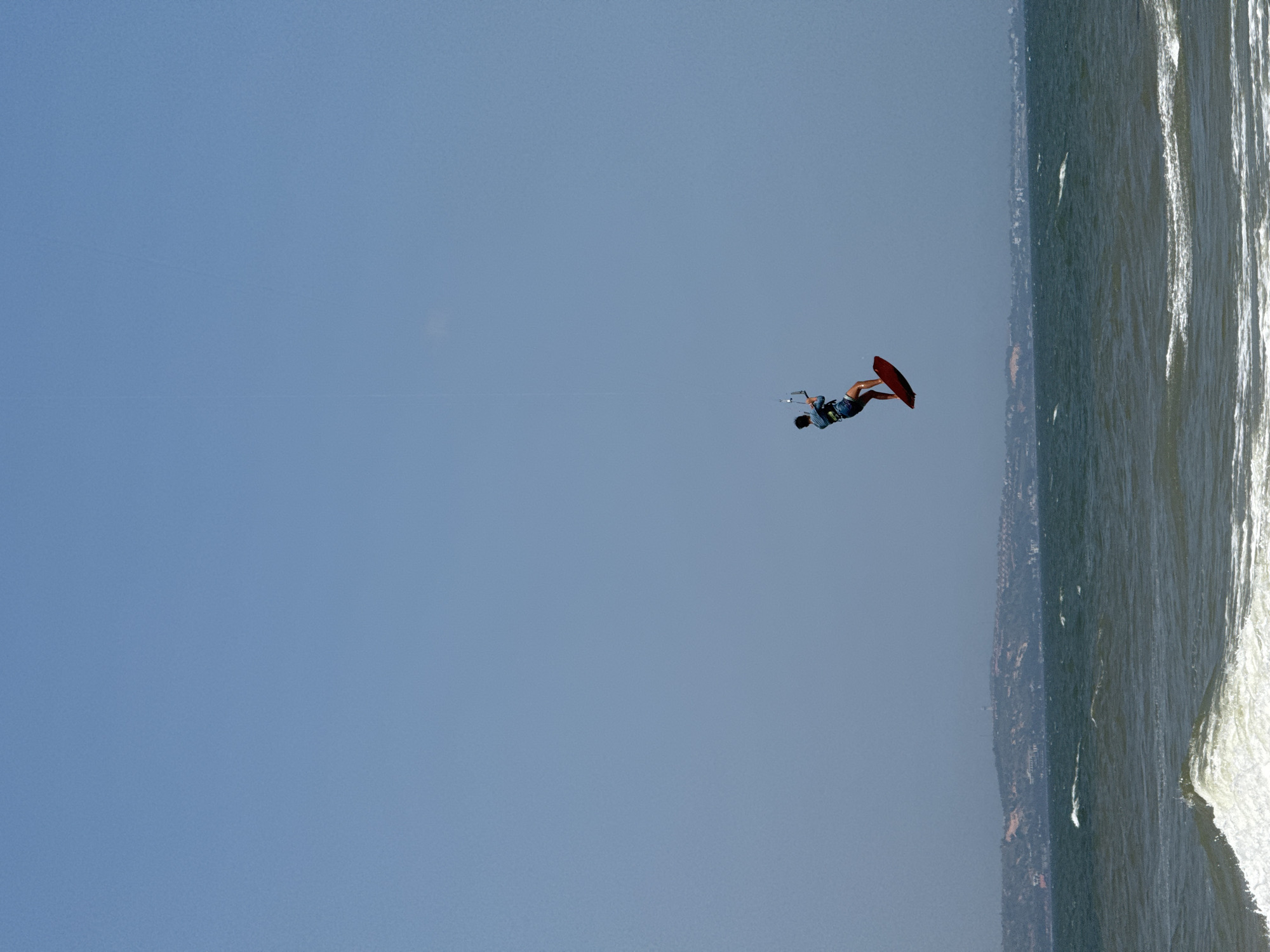 A kitesurfer is captured mid-air against a clear blue sky, performing a trick over the ocean in Mũi Né, Vietnam. The kitesurfer, wearing a blue and white striped shirt and denim shorts, is positioned to the left of the frame, riding a red kiteboard. Their body is contorted as they hold onto the control bar, suggesting an aerial maneuver. The kitesurf kite, a white canopy, is visible above and slightly to the right of the kitesurfer, connected by lines. Below the kitesurfer, the dark green water of the sea is choppy with white crests on the waves, indicating wind. In the distance, a hazy coastline with rolling hills and scattered buildings stretches across the horizon. The sky is a uniform pale blue, with no clouds visible. The overall scene conveys action and the exhilaration of watersports in a coastal environment.