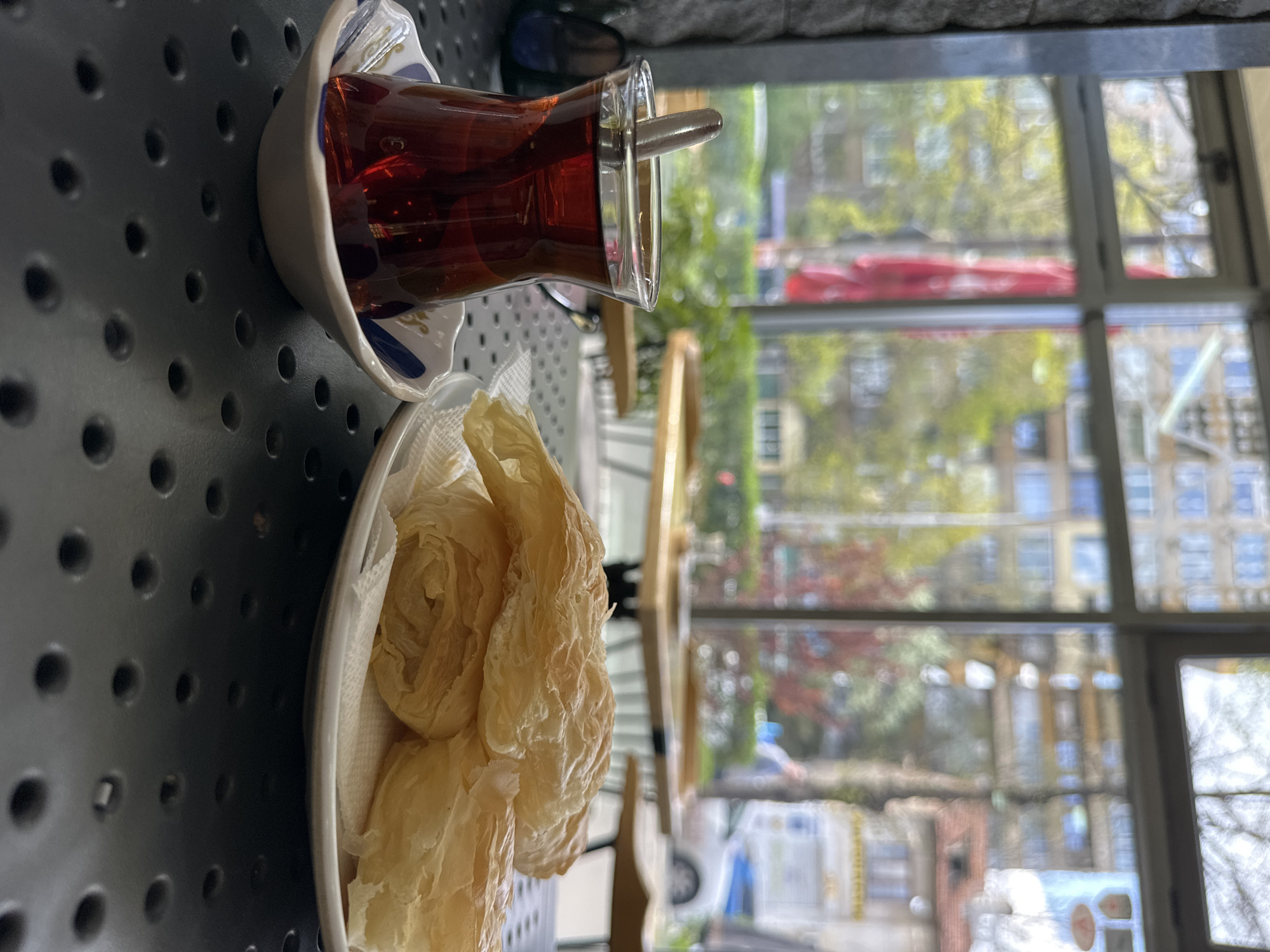 This vertical image depicts a close-up of a table setting inside what appears to be a cafe or restaurant in Sofia, Bulgaria, with an urban outdoor scene visible through a large window.
The primary subjects are a glass of dark tea and a plate of pastries. The tea is served in a traditional tulip-shaped glass, containing a small spoon, and rests on a white saucer with blue and gold decorative patterns. Adjacent to the tea is a white plate holding several pieces of light-colored, flaky pastry, characteristic of phyllo dough pastries like banitsa, a popular Bulgarian dish. These pastries appear delicate and layered. The table surface is dark grey and features a distinct perforated pattern of small holes.
Through the window in the background, a blurred outdoor environment is visible. This includes green foliage from trees, indistinct building facades, and a prominent red structure or awning in the middle distance. A blurred white vehicle is also faintly visible on the street.
The natural light streaming through the window indicates it is daytime, likely morning or early afternoon. No people are visible in the frame, and the scene evokes a sense of quiet relaxation, suggesting a moment for a beverage and snack. There is no visible text in the image.