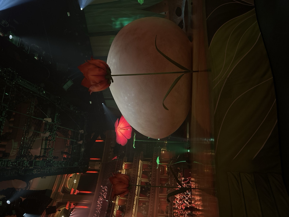 A theatrical set features a large, pale sphere resembling a moon or egg, adorned with an oversized flower. Below, a crowd of people fills a tiered seating area, with complex scaffolding and architectural details forming the backdrop. Additional illuminated flower props add to the surreal ambiance. A leafy panel looms to the right, partially obscuring the scene. The lighting and composition evoke a dreamlike or fantastical setting.