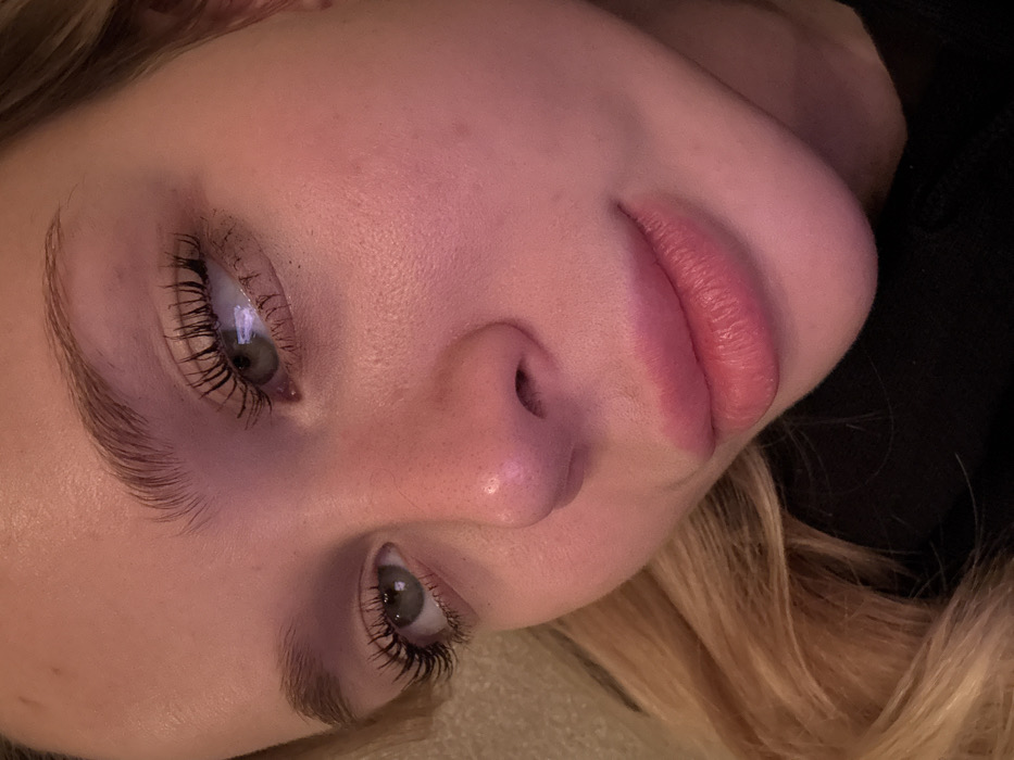 A close-up, angled portrait of a fair-skinned person with striking features. They have prominent eyelashes coated in mascara, defined eyebrows, and full lips. The skin appears smooth, possibly due to makeup or lighting. One side of the face is partially obscured by dark fabric, while the other is framed by blonde hair. The overall tone is soft and romantic, accentuated by the pinkish lighting.