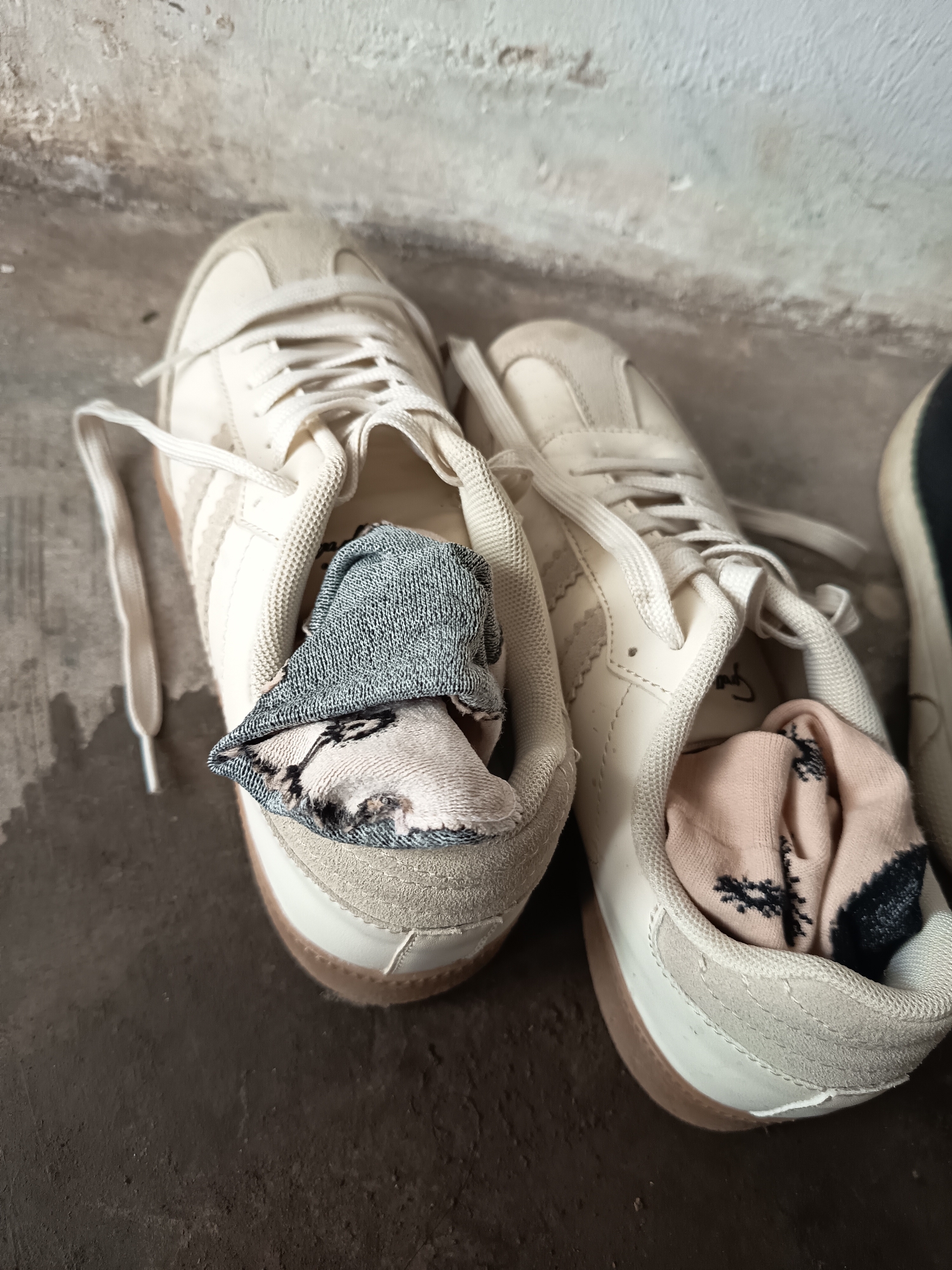 Two white sneakers with brown soles are on a concrete floor next to a textured wall. The left sneaker contains a rolled-up grey sock with a patterned interior visible. The right sneaker contains a rolled-up pink sock with a black abstract pattern. The shoes are laced with white laces. A portion of a dark shoe is visible to the right of the white sneakers. The scene is located in Krampilan, Indonesia.