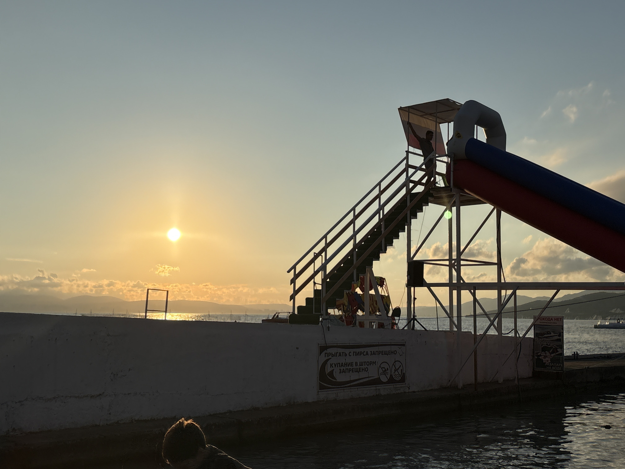 The image depicts a sunset over the Black Sea, likely at a recreational area in Novorossiysk, Russia. A silhouetted person stands at the top of a wooden staircase leading to an inflatable water slide, which is red and blue. The sun is low on the horizon, casting a warm golden glow on the sky and clouds, and reflecting on the calm water. In the background, distant hills and boats are visible. A white concrete wall runs along the edge of the water, with several signs attached to it. One sign, in Russian, reads "JUMPING FROM THE PIER IS PROHIBITED" and "SWIMMING IN ROUGH SEAS IS PROHIBITED." Another sign, partially visible, shows images related to water activities. In the foreground, another silhouetted person is partially visible, looking towards the water. The scene suggests a tranquil end to a day at the seaside.