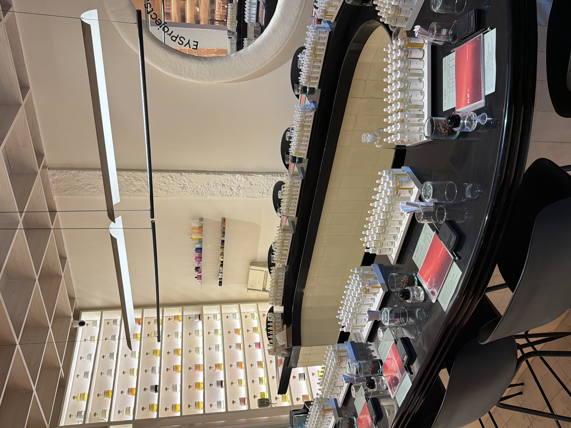 The image depicts an indoor perfume workshop or laboratory setting, possibly located in a city like Mengwi, Indonesia, given the context. The foreground features a dark, curved counter with multiple small glass bottles of perfume oils or essences arranged on white trays. Some bottles are clear, revealing light-colored liquids, while others appear to contain darker hues. Several small notebooks with red covers and green pages are laid out on the counter, alongside small digital scales and glass beakers. Two empty bar stools with black seats and thin metal legs are visible at the counter.
In the mid-ground, a curved, cream-colored counter with a black top extends across the scene, also holding trays of perfume bottles and small glasses. Behind this counter, a white shelving unit is filled with numerous identical glass bottles, each containing a different colored liquid, arranged in neat rows. On a high shelf in the background, a collection of colorful small bottles is displayed.
The environment is brightly lit by overhead fluorescent lights. A large, circular mirror with a textured border is mounted on the wall in the background, reflecting parts of the room. The walls are white, with one section featuring a textured finish. The ceiling has a grid pattern composed of thin wooden slats. There are no people visible in the image. The overall impression is one of a well-organized and equipped space dedicated to the art and science of perfumery.