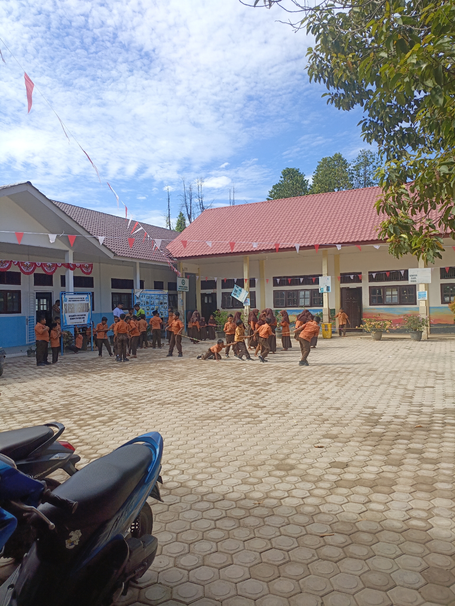 The image captures an outdoor scene in front of a school building, likely a primary school given the age of the children. Several children, dressed in orange shirts and brown pants, are engaged in a game of tug-of-war on a paved courtyard. The courtyard is made of hexagonal concrete tiles. The school building has a red tiled roof and light-colored walls. Red and white triangular flags are strung across the facade of the building and also hang from a wire overhead, suggesting a celebration or national event. Some children are in the process of pulling a rope, while others are observing. The sky is blue with scattered white clouds. Two motorbikes are parked in the foreground, one blue and one dark-colored. The environment appears to be a schoolyard in a warm, sunny climate.