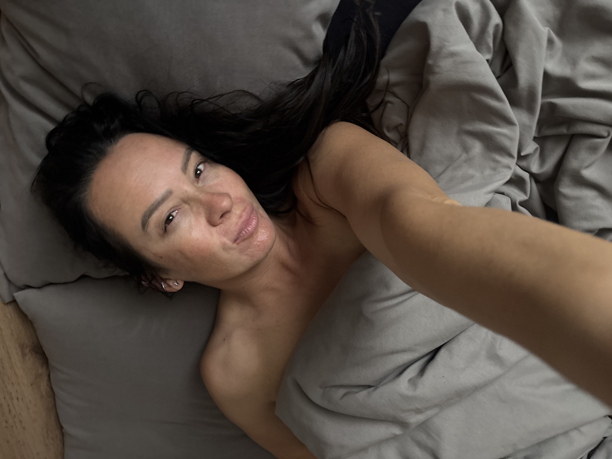 A woman lies in a bed with grey sheets and a grey duvet. She has dark hair and is partially covered by the duvet. Her right arm is extended towards the camera, suggesting she is taking a selfie. The location is indeterminate; there is no visible evidence indicating the city of Chaikovsky, Russia.