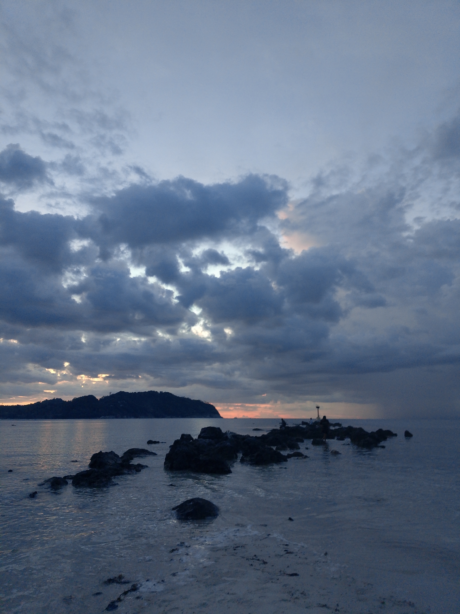 This image depicts a coastal scene at dusk or dawn near Ko Pha Ngan, Thailand. The sky is filled with dramatic, dark clouds, with patches of soft, warm light peeking through, suggesting the sun is either rising or setting. In the distance, a silhouetted island or peninsula, densely covered in vegetation, forms the horizon line. In the foreground, dark, jagged rocks emerge from the calm, dark water, creating a path-like formation extending towards the viewer from the center of the frame. Several small figures, appearing to be people, are visible standing on these rocks, some appearing to be interacting. The water is shallow near the shore, revealing a sandy or rocky bottom with some scattered debris. The overall mood is serene and somewhat moody due to the overcast sky and low light.