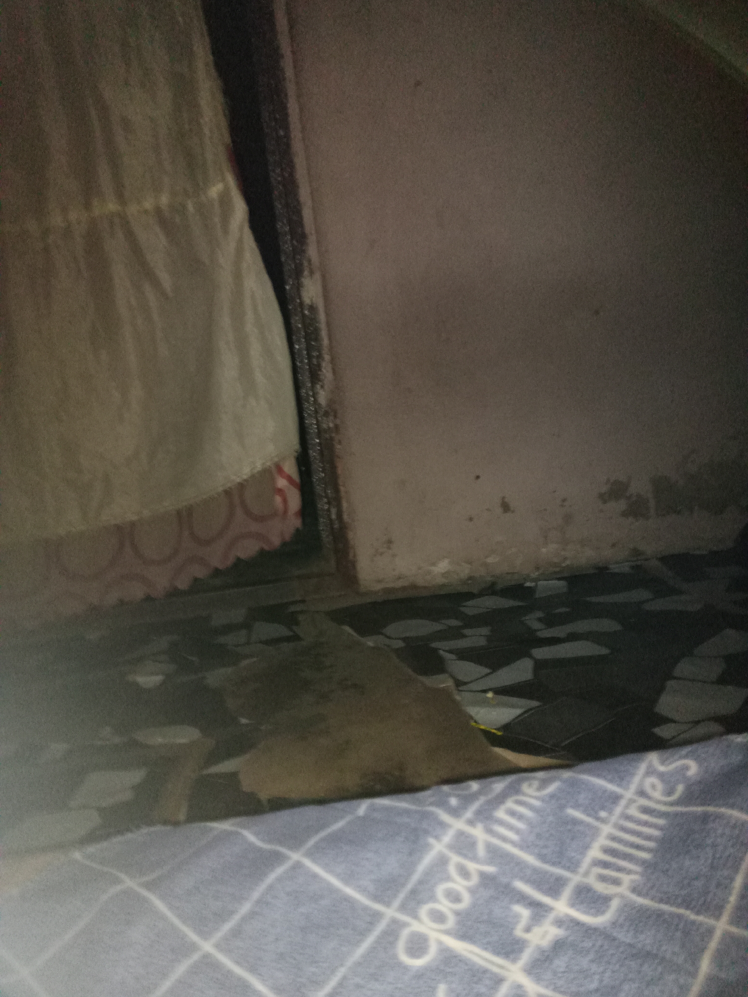 The image depicts an indoor scene with a tiled floor and a wall. A light-colored fabric, possibly a curtain or bedding, hangs from the left side of the frame, with a patterned fabric visible underneath. A dark, flat object is partially visible on the floor in the center of the image. The wall on the right appears to be textured and discolored in some areas. The lower portion of the image shows a blue fabric with white text and grid patterns. The scene is dimly lit.