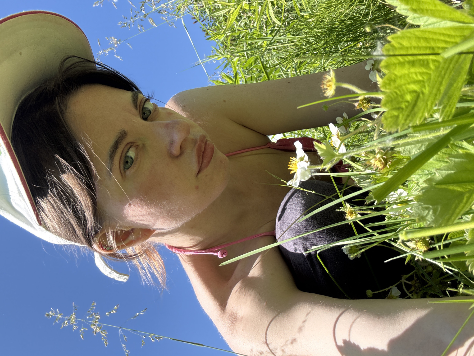 The image shows a young woman from a low-angle perspective, crouching in a field of green grass and small white flowers under a clear blue sky. She is wearing a white baseball cap, a pink bikini top, and dark pants. The woman is looking directly at the camera with a neutral expression. The background consists of a bright blue sky and tall blades of grass and wildflowers. There is no visible indication of the city of Chaikovsky, Russia, in the image.