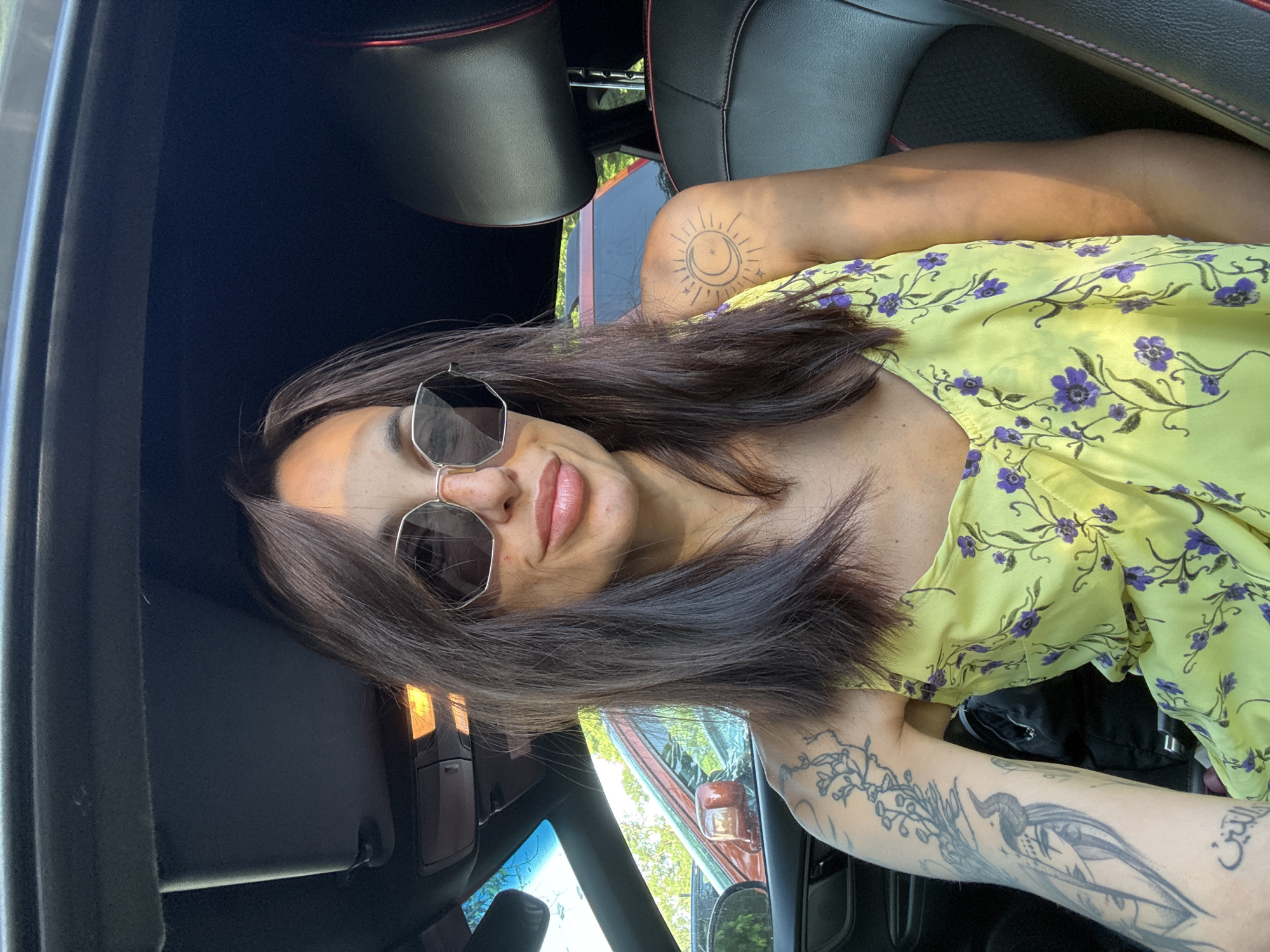 A woman with long, dark hair, wearing a yellow floral dress and sunglasses, is seated in the passenger seat of a car. The car's interior is visible, including the black leather seat and headrest. Outside the car, through the window, another car and trees can be seen. The image appears to be taken during the daytime. There is no indication that the image was taken in Pleven, Bulgaria.