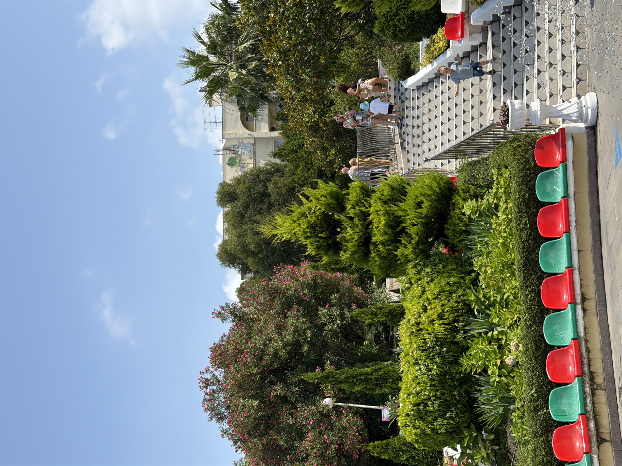 The image depicts a sunlit outdoor scene with a focus on a grand staircase adorned with geometric tile patterns, ascending into a lush green environment. Several people are present on the stairs and in the surrounding area, engaged in various activities. A young boy in a gray t-shirt and blue shorts is running down the stairs. Further up, a family group is seen ascending, including a woman in a colorful floral dress and sunglasses, another woman in a black top and beige shorts, and children. A decorative white planter with a plant sits at the base of the stairs.
In the foreground, a row of vibrant red and green plastic stadium seats is visible, suggesting a public space or performance area. The background is filled with abundant vegetation, including flowering trees with pink blossoms, various evergreen shrubs and trees, and palm trees. A building with a light-colored facade and some architectural details is partially visible behind the trees. The sky is a clear blue with a few wispy clouds. The overall atmosphere is one of a pleasant, sunny day in a well-maintained outdoor recreational area. This location appears to be a public park or garden within the city of Sochi, Russia, likely designed for leisure and viewing.