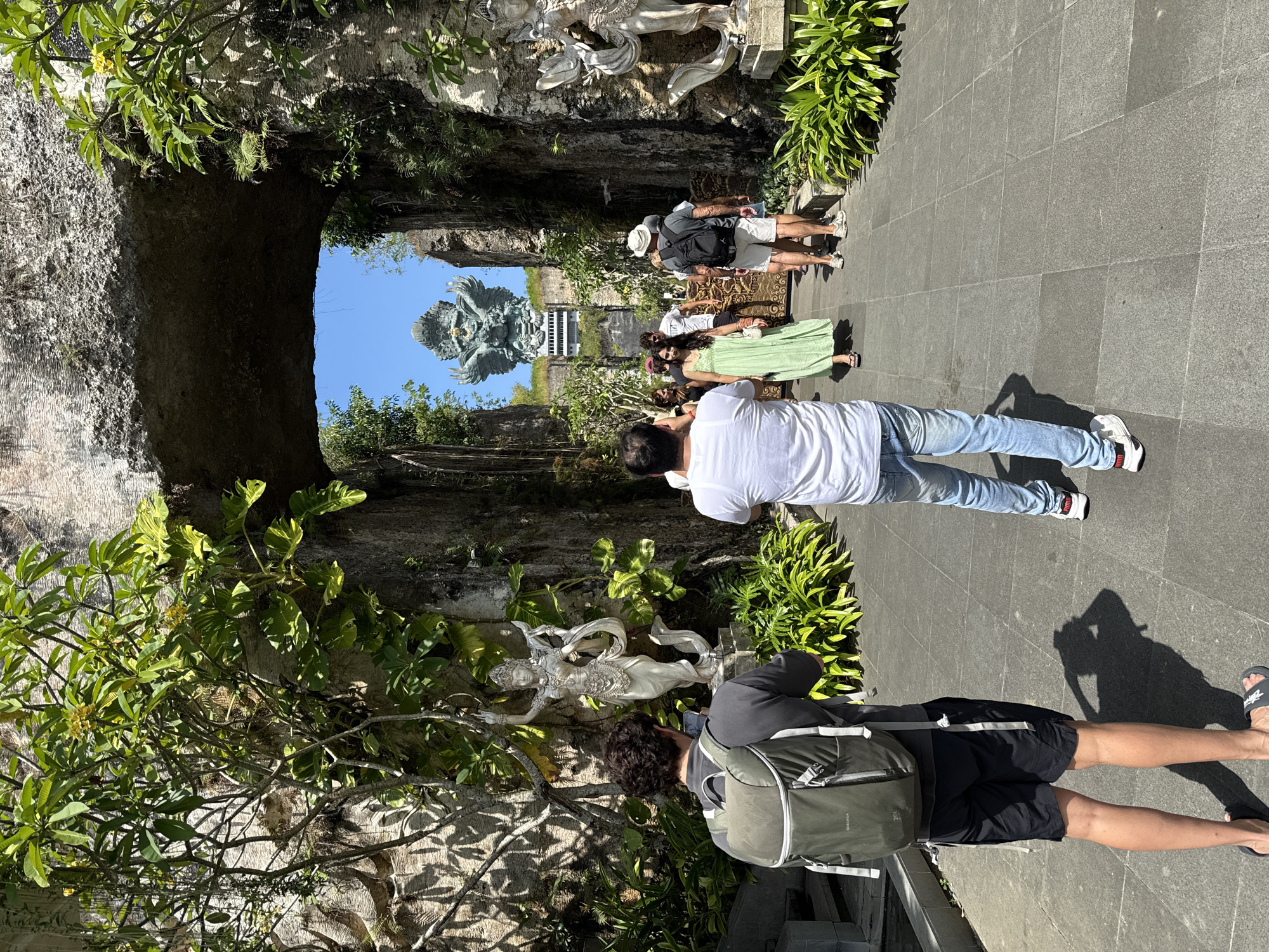 The image shows a view through an archway in Kuta Selatan, Indonesia, towards a large Garuda Wisnu Kencana statue. Several tourists are present, including one man in a white shirt and jeans who appears to be taking a photo, and another man in a dark shirt and shorts with a backpack who is also taking a photo of a white sculpture. The archway is made of stone and is surrounded by lush greenery. The ground is paved with grey tiles.