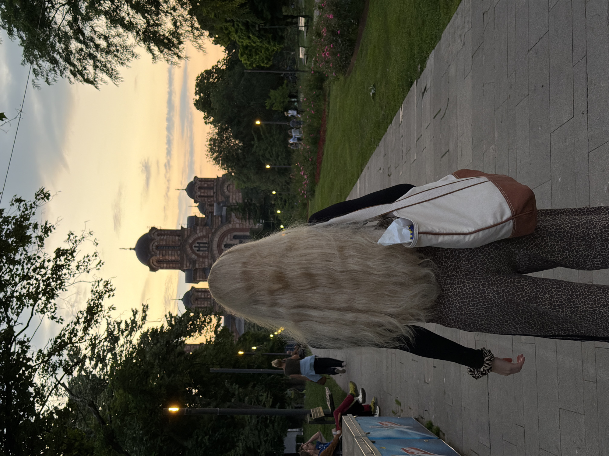 A sideways-oriented photograph shows a woman with long, blonde hair, walking along a paved path. She is wearing patterned pants and a black top, carrying a tote bag. In the background, a multi-domed Orthodox church is visible against a sky with an orange and yellow sunset. The scene appears to be a park or urban area, possibly in Vracar Urban Municipality, Serbia, given the architectural style of the church. People are seen on benches and the path. Trees and grass line the path, and the lighting suggests it is late afternoon or evening.
