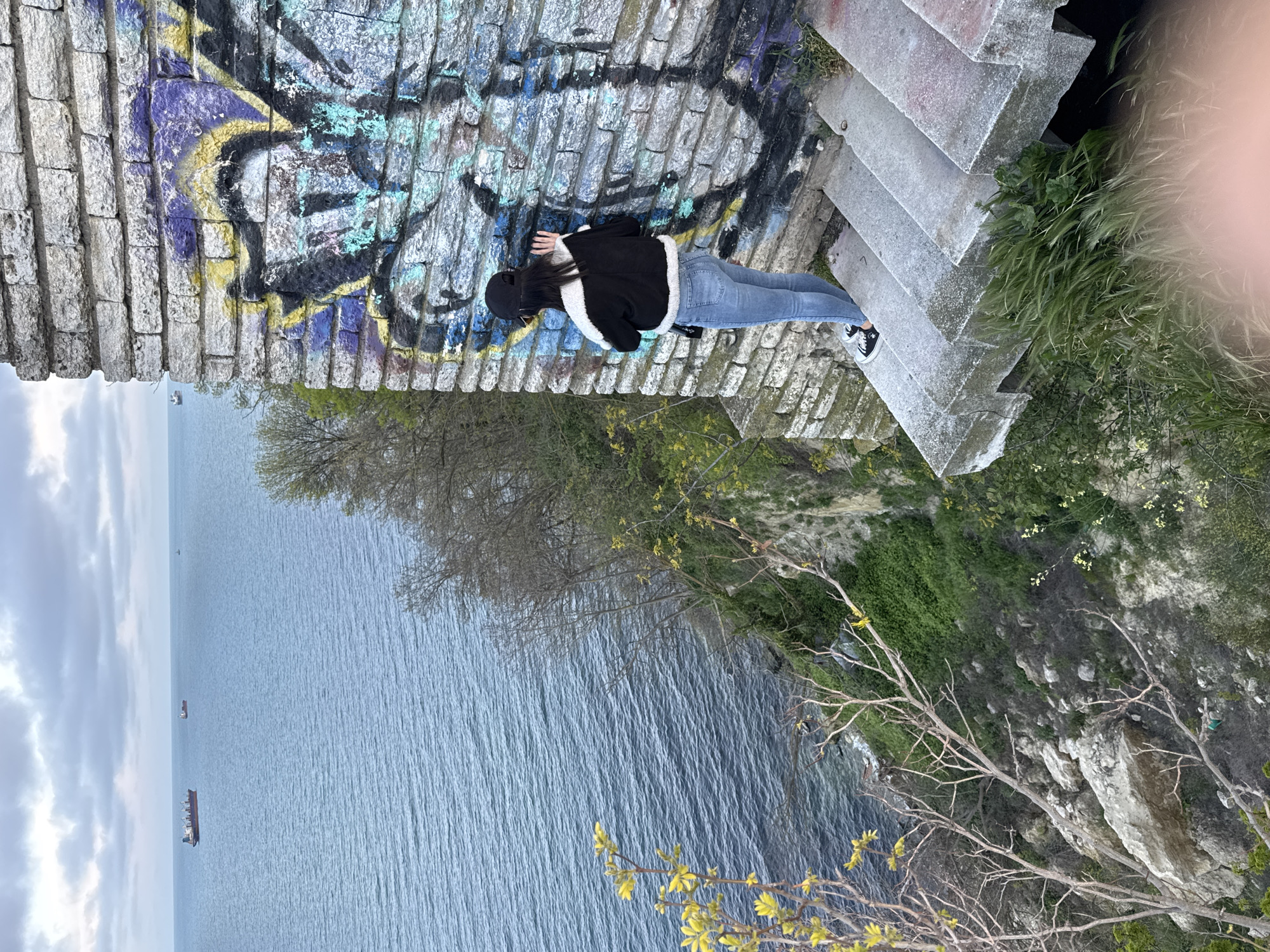 A rotated, medium shot captures a scene on the coastline of Varna, Bulgaria. A woman in a black cap, black jacket with white fur trim, and blue jeans lies prone on a stone wall adorned with graffiti. The wall is textured, consisting of stacked stone blocks. To the right of the woman, concrete steps descend. Below the wall and steps, a cliff covered with green vegetation leads down to the blue sea. In the distance on the sea, there are ships. The sky is cloudy. The image appears to have been taken from above. A hint of a pink object (likely part of the photographer's hand) is visible in the top right corner.