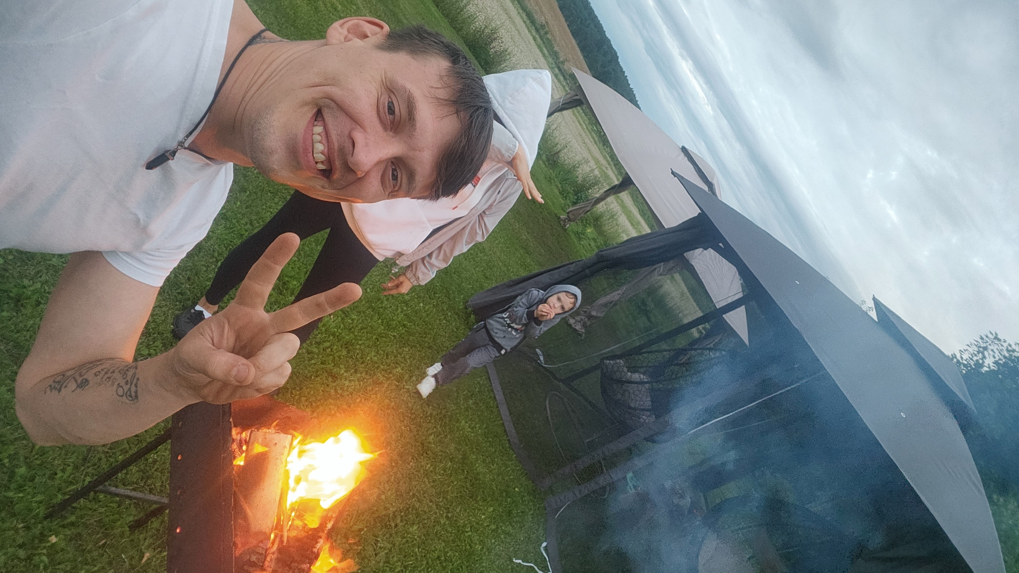In Ezernieki, Latvia, a man with dark hair and a white t-shirt smiles at the camera, making a peace sign with his right hand. He has a tattoo on his right arm. Behind him, a person in a white hoodie and dark pants is partially visible, with their arm extended. Further back, a child in a grey hoodie and dark pants stands near a black gazebo. A fire burns in a metal brazier to the right of the man. The scene is set outdoors on a grassy area with a field and trees in the background under a cloudy sky.