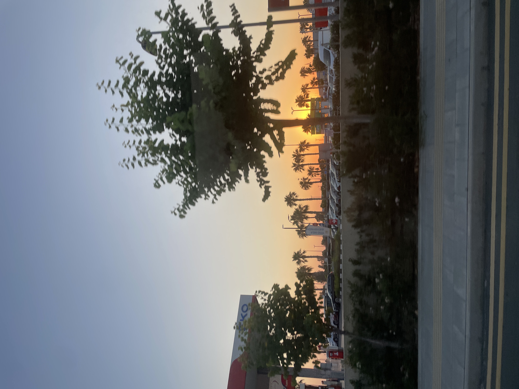 The image captures a sunset scene at a gas station, likely in Timi Community, Cyprus, given the presence of palm trees and the style of architecture. In the foreground, a road with yellow markings and a grey sidewalk is visible. Behind the sidewalk, a line of dark green bushes separates the road from a parking lot filled with various cars, including sedans and vans. A white van is parked prominently in the lot.
The gas station itself, branded with "TEKO" and a red stripe, stands to the left of the frame, with a red structure, possibly a fuel pump, visible in front of it. Several palm trees are scattered throughout the scene, silhouetted against the vibrant orange and yellow hues of the setting sun. The sky transitions from a pale blue at the top to warm tones closer to the horizon. Two trees with green foliage are in the mid-ground, one on the left and another larger, more prominent tree on the right, partially obscuring the view of the sun. Streetlights are also visible, some illuminated. A tall pole with signage in Greek, possibly indicating directions or business names, stands near the palm trees on the right. The overall impression is one of a typical evening at a commercial establishment in a warm climate.