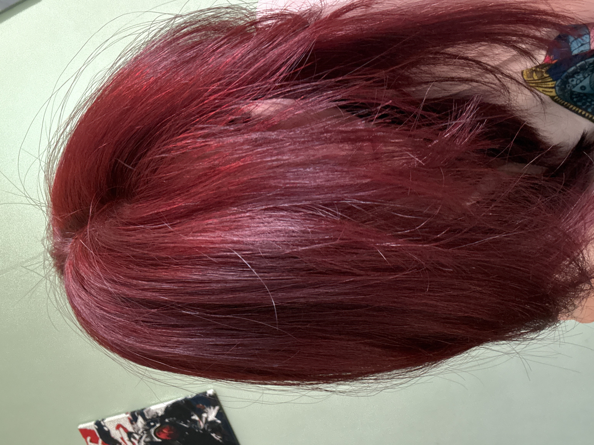 A close-up, waist-up side profile shot captures a person with vibrant, dark red hair styled in choppy layers. The hair is the dominant feature, with some strands appearing lighter and shinier, suggesting a recent dye job. A hint of a colorful patterned garment is visible at the bottom right of the frame. In the upper left corner, a portion of a framed artwork, featuring a dark, graphic design with red accents, hangs on a light green wall. The lighting is even, casting soft shadows and highlighting the texture of the hair. The background is a plain, pale green wall. There are no other discernible objects or people in the frame. The image appears to have been taken indoors, possibly in a residential setting.