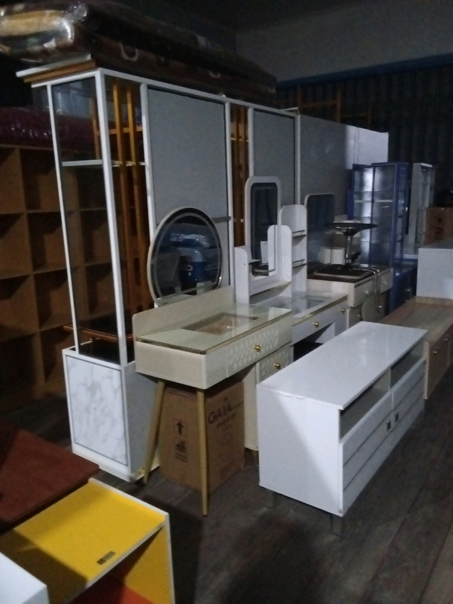 The image depicts an indoor scene, likely a warehouse or store, filled with various pieces of furniture. In the foreground, a white dresser-like cabinet with multiple drawers and a light wood top is prominently displayed. To the left of this, a small, bright yellow table with an open compartment is visible. Further back and to the left, a tall, white-framed cabinet with glass doors and wooden shelves stands against a background of wooden shelving units.
Dominating the center of the image is a vanity table with a mirrored top and a light-colored countertop. It has drawers with gold-toned knobs. The vanity is supported by wooden legs and sits atop a cardboard box with a "GAIA" label.
To the right of the vanity, a blue glass-fronted cabinet is partially visible. Behind the vanity, there are more furniture pieces, including what appears to be a desk with a chair and other cabinets. The overall environment is dimly lit, with a dark, possibly concrete or tiled floor. There are no people visible in the image. The context of "Gas Alam Badak I, Indonesia" suggests this might be a retail or storage space within that location, though the image itself doesn't provide explicit geographical markers.