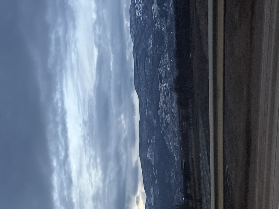 A landscape features a mountain range under a cloudy sky. Light breaks through the clouds towards the bottom. The mountain is dark with hints of snow on its peaks and ridges. The foreground suggests a view from inside a vehicle, with a window frame visible. The image orientation is vertical.