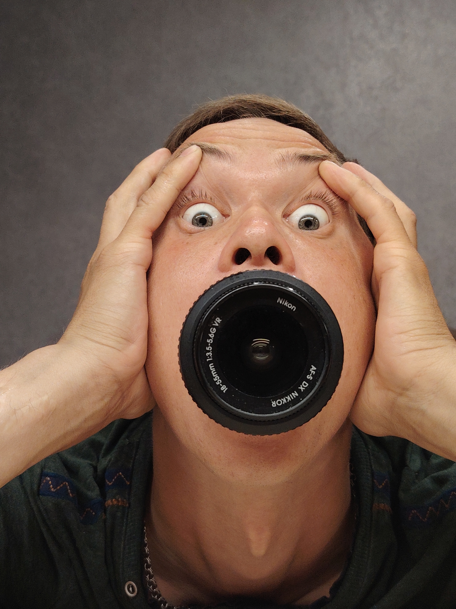 The image is a close-up portrait of a person's face, centered and filling most of the frame. The person's eyes are wide open, with a look of surprise or shock. Their hands are raised, gripping their temples, further emphasizing the expression. A camera lens is inserted into the person's mouth, obscuring it. The lens is black and has white lettering indicating it is a Nikon lens, model AF-S DX NIKKOR 18-55mm 1:3.5-5.6G VR. The background is a plain, dark gray, textured surface, possibly a wall or backdrop. The lighting appears to be artificial, creating some shadows on the face. The person is wearing a dark green shirt with a patterned design on the shoulder area. There is no discernible location in the city of Chaikovsky, Russia, or any other specific geographical markers in the image.