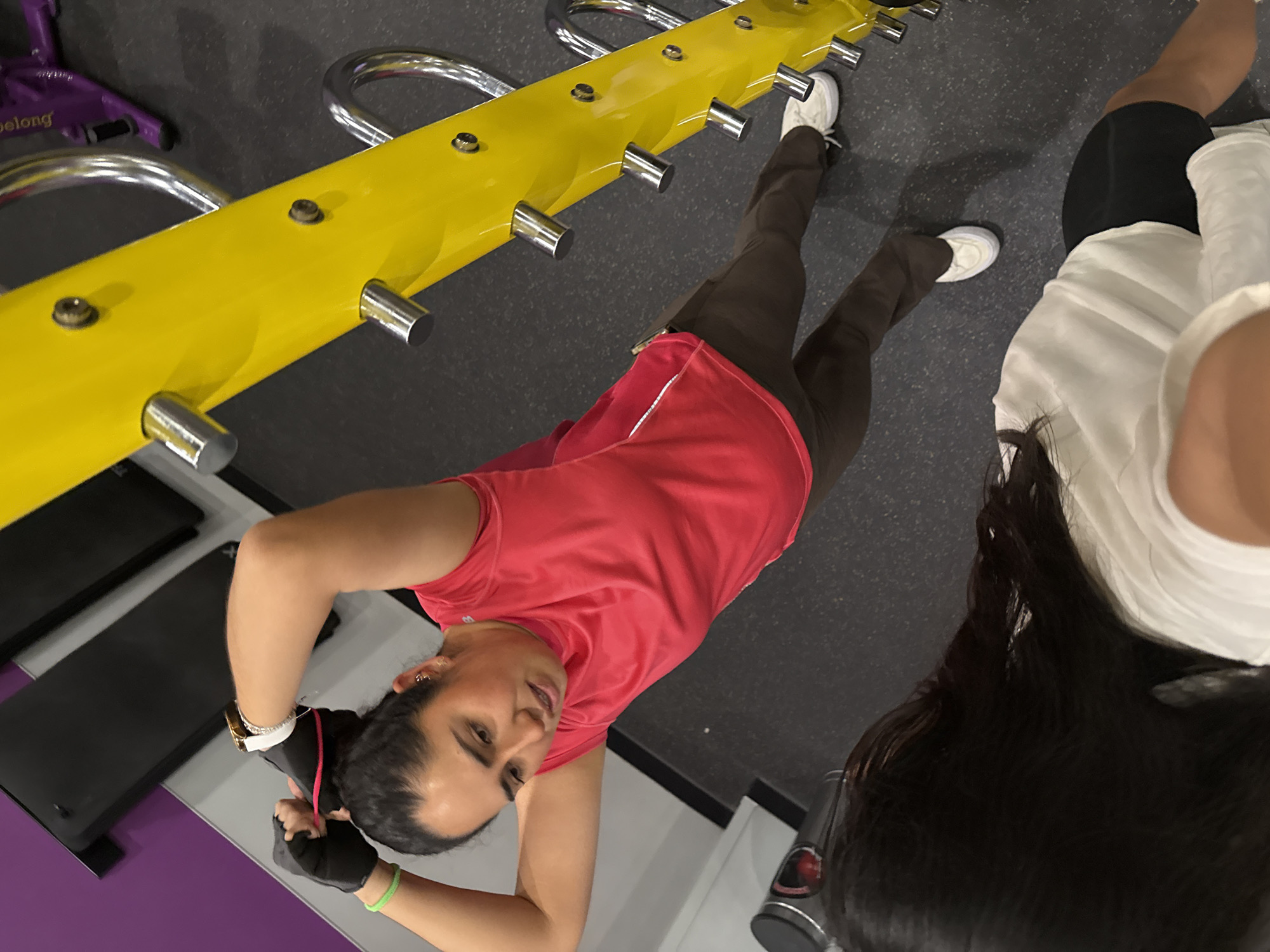 The image depicts two individuals at a fitness center, likely Planet Fitness, given the distinctive purple and yellow equipment visible in the frame. One person, likely the photographer or subject of the photo, is positioned in the lower portion of the image. They are wearing a red shirt and brown athletic pants, as well as fitness gloves on their hands. The individual's head is tilted downward towards the camera, making eye contact. The other person, in the upper right of the image, is wearing a white shirt and black athletic shorts. Her hair is long, dark, and cascades down her back. A piece of yellow fitness equipment, featuring several silver cylindrical attachments, is positioned horizontally across the upper portion of the image. In the top right corner, there's a pair of athletic shoes. The flooring appears to be a dark grey rubberized material, typical in gyms. The location is a Planet Fitness in the city of Puebla, Mexico.