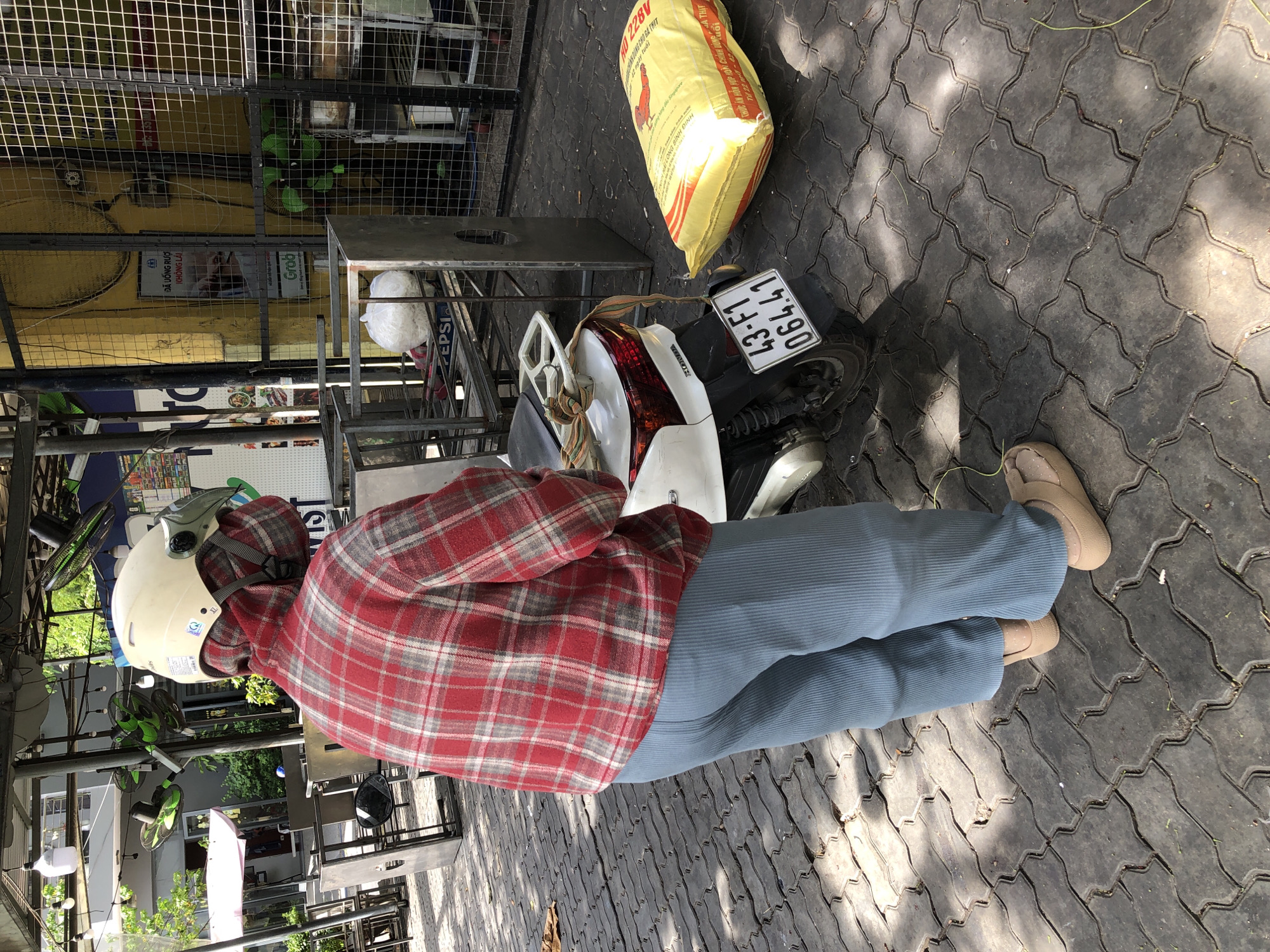 A person is standing next to a white scooter parked on a paved area. The person is wearing a red and grey plaid shirt, light blue pants, a white helmet, and tan slip-on shoes. The scooter has a license plate that reads "43-F1 064.41". A large yellow bag is lying on the ground next to the scooter. The background shows a covered outdoor area with tables and chairs, and a metal cage structure. This scene appears to be in Đà Nẵng, Vietnam, likely on a street or in a public area.