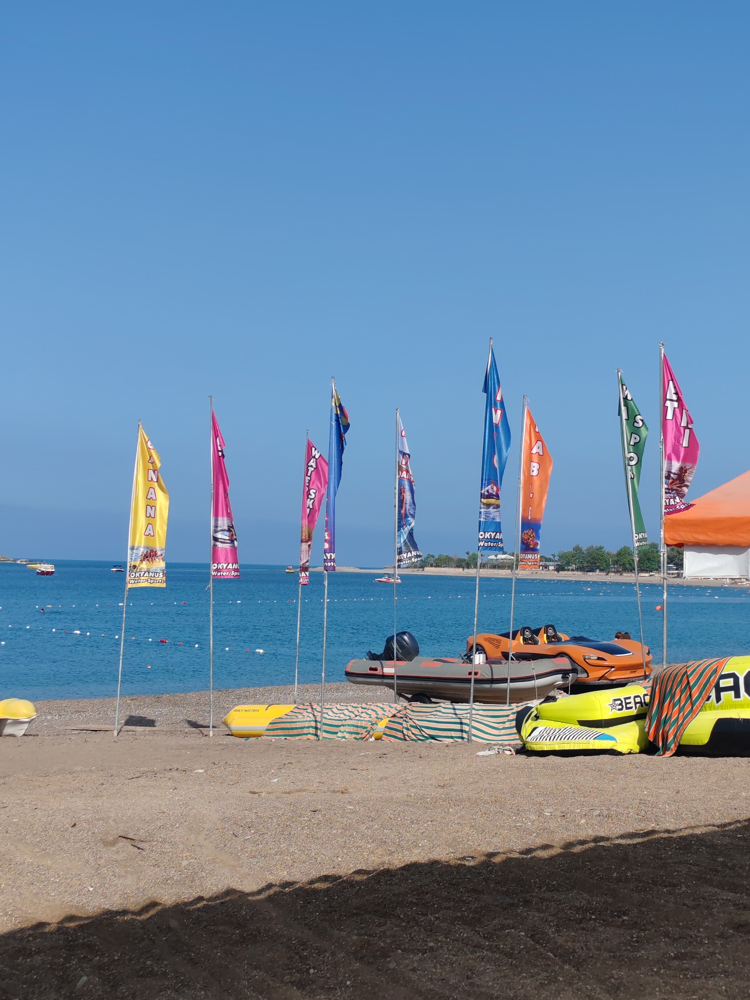 The image captures a sunny day at a beach in Alanya, Turkey. The clear blue sky and calm turquoise sea indicate favorable weather conditions. A row of colorful advertising flags, promoting water sports activities under the brand "OKYANUS Water Sports," stands on the sandy beach. Various watercraft are parked near the flags, including an orange jet-ski-like vehicle, a gray inflatable boat with a motor, and yellow inflatable watercraft. The beach itself is composed of sand and small pebbles, with some parts shaded. In the background, the coastline is visible, along with boats at sea. The scene portrays a typical beach setup for water sports activities in the coastal city of Alanya.