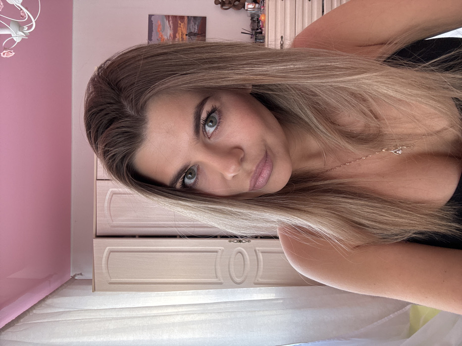 An indoor photograph features a single adult female, appearing to be in her 20s or 30s, positioned centrally and facing the camera directly. She has long, straight, multi-toned blonde and light brown hair with a middle part, green eyes, and light-toned skin. Her lips are slightly parted and adorned with light pink lipstick. She is wearing a dark, possibly black, strapless garment, with a delicate gold-colored chain necklace and a small teardrop-shaped pendant visible on her décolletage.
The background consists of a residential interior. The ceiling is painted a solid bright pink, with a partial view of a white, decorative, rose-embellished ceiling light fixture in the upper right. To the viewer's left, a sheer, white curtain hangs, partially obscuring a light-colored wooden wardrobe or cabinet featuring two paneled doors. To the right, a light-colored wooden chest of drawers or dresser is visible, displaying several objects including a dark, abstract sculpture (possibly two intertwined figures), a clear container holding brushes or pens, and other indistinct items. Above the dresser, a framed painting depicting a sunset or sunrise over a body of water with a solitary figure walking on a beach is mounted on a light-colored wall. The scene is well-lit, suggesting diffused natural and artificial light. The specific location within the city of Chaikovsky, Russia, cannot be confirmed from the visual information present in the image.