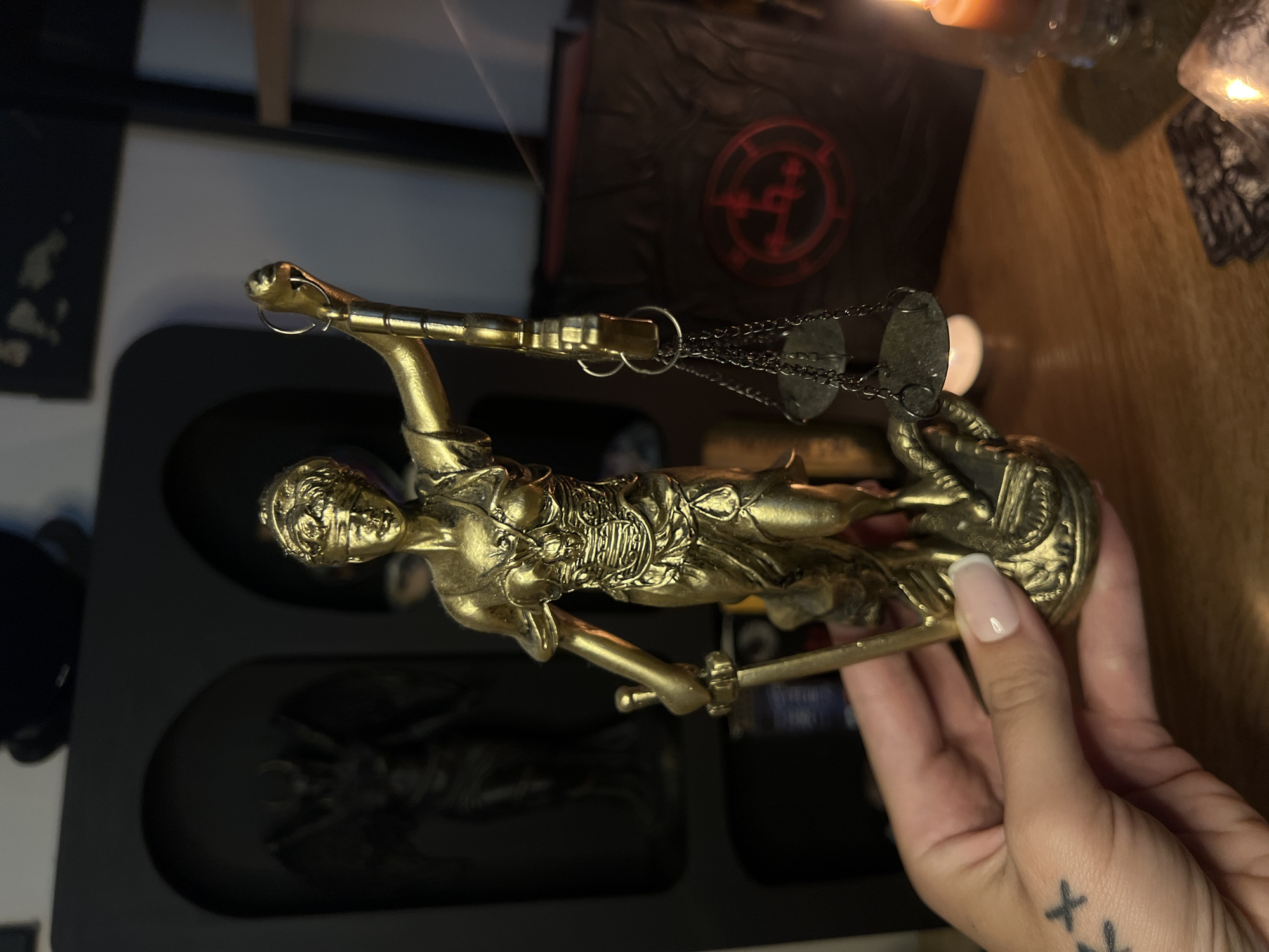 A gold-colored statue of Lady Justice, a blindfolded female figure holding a sword and scales, is held in a person's left hand. The statue is detailed, with the figure wearing a toga and armor, and a serpent coiled around the base. In the background, there is a dark shelf with a dark figurine and a book with a red symbol on its cover. A lit orange candle is visible on the right side of the image, and a deck of cards is on the table in the foreground. The scene is set indoors, likely on a wooden table.