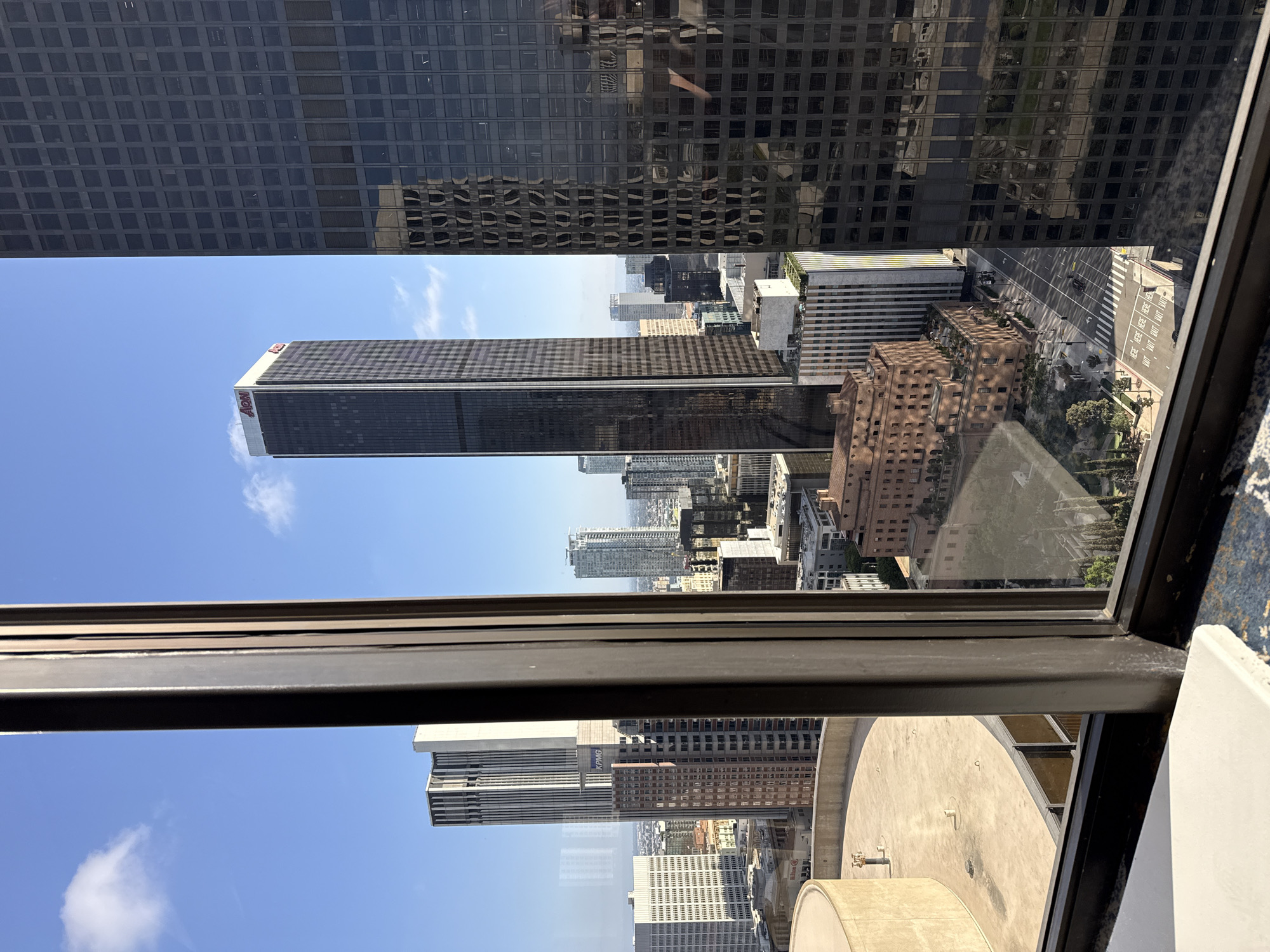The image is an exterior, elevated view looking through a window at the Los Angeles skyline. The time appears to be daytime, with a clear blue sky and scattered clouds. In the foreground, a dark-colored window frame runs vertically down the center of the image, dividing the scene.
Dominating the mid-ground and background are numerous skyscrapers of varying architectural styles and colors. To the left of the window frame, a beige, circular building with a flat roof and railing is visible. Further back, a tall, slender skyscraper with a white top and the red "AON" logo stands out. To the right of the window frame, a large, dark, rectangular skyscraper with a grid of windows fills a significant portion of the frame. Its reflective surface mirrors other buildings in the skyline.
In the lower portion of the image, a street scene is visible, with cars driving and pedestrians walking. Traffic lights and crosswalk markings are present. Several other buildings of different heights and materials are scattered throughout the urban landscape. Trees and some greenery are also present, particularly in the lower left and right sections of the image. The overall impression is a dense urban environment.