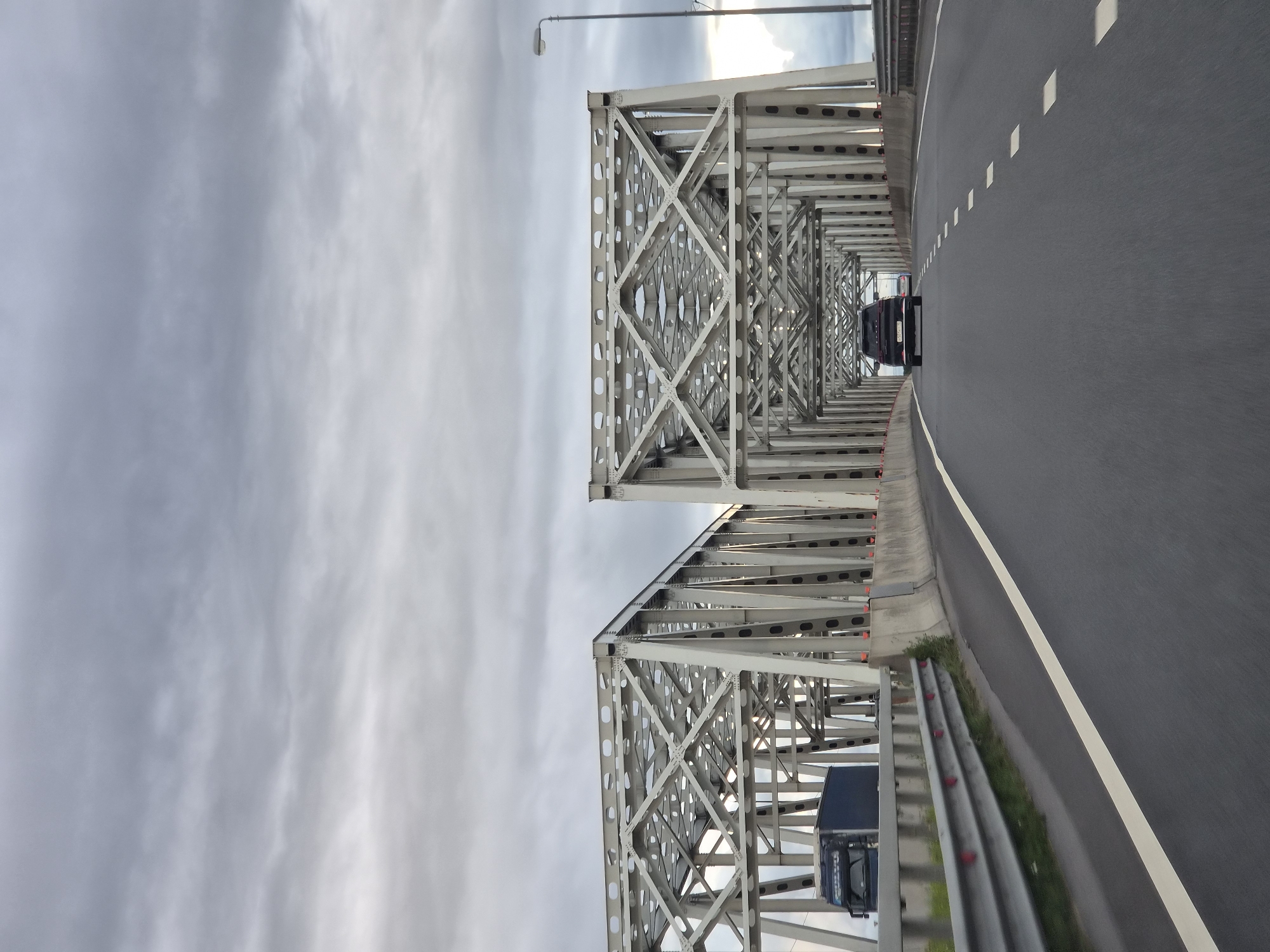 An outdoor, eye-level, medium shot captures a view from a vehicle driving across a bridge in Yushkovo, Russia. The bridge, constructed from grey metal trussing, spans over an unseen body of water. The sky above is overcast with grey clouds. On the bridge, a dark-colored SUV is driving in the right lane, followed by a white car. In the left lane, a dark blue truck with a blue cargo box is traveling in the opposite direction. The road surface is asphalt with visible white lane markings. A grey metal guardrail runs along the left side of the road, separating it from the embankment. The scene depicts typical highway traffic on a cloudy day.