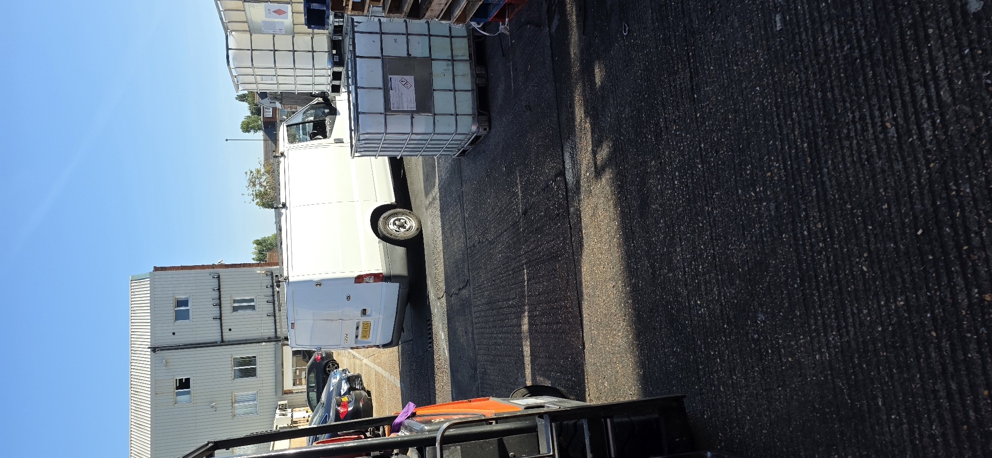 An outdoor, daytime scene depicts an industrial or commercial setting under a clear blue sky. In the foreground, the edge of an orange forklift is visible on the left. Beyond the forklift, a white Ford Transit van is parked on an asphalt surface. Several dark-colored cars are parked in a lot to the left of the van. In the background, a light-colored building with multiple windows and a corrugated roof is situated. To the right of the van, two large, white IBC (Intermediate Bulk Container) totes are stacked. The ground is a textured asphalt surface, with distinct lines or grooves visible. Some shadows are cast across the asphalt, indicating bright sunlight. The scene appears to be in a location such as Arun, United Kingdom, given the context.