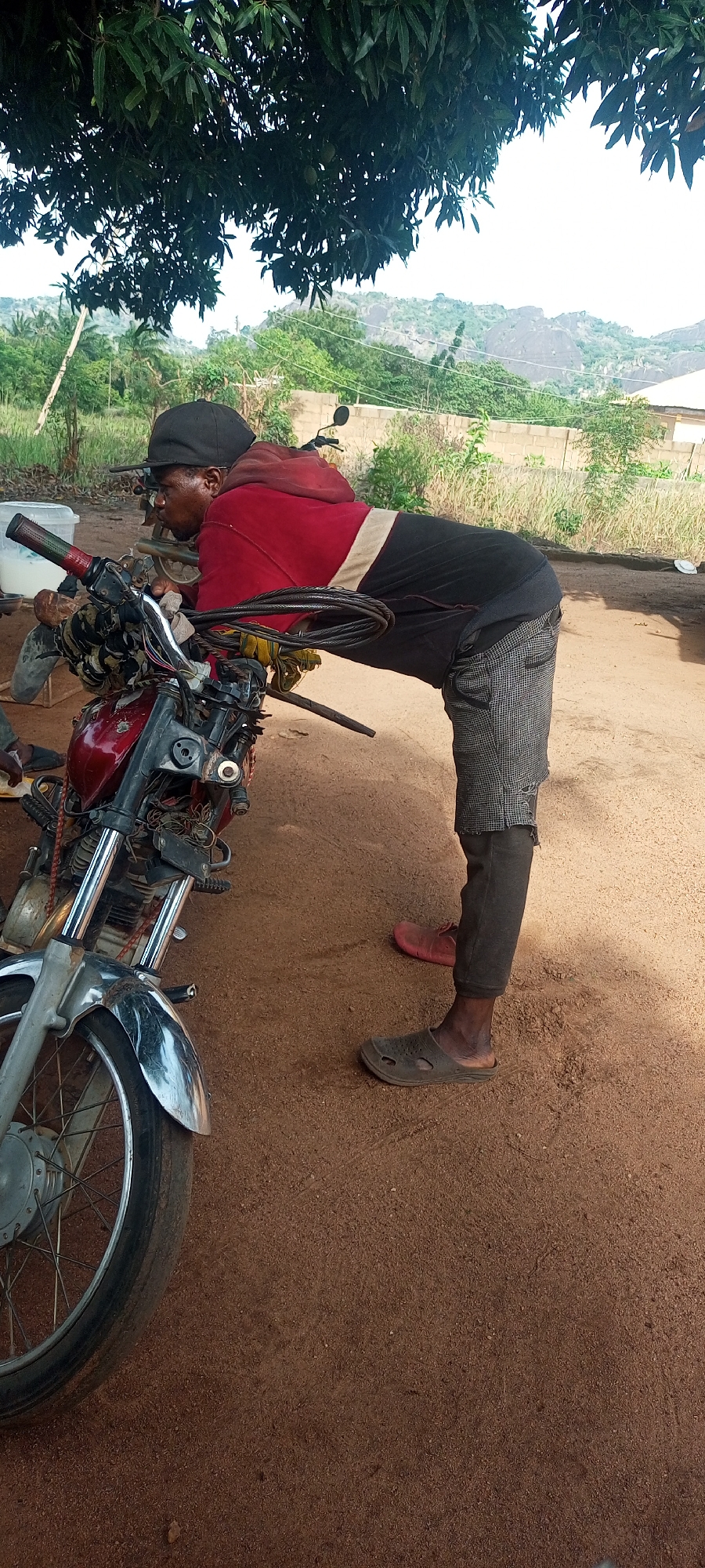 An outdoor, eye-level shot captures a man leaning over a red motorcycle on a dirt ground, under the partial shade of a large tree. The man, positioned on the right side of the frame, is wearing a black baseball cap, a red hooded sweatshirt over a black t-shirt, patterned shorts over dark pants, and dark gray crocs on his feet. One red shoe is also visible in the background near his left foot. He is bent forward, resting his upper body and arms on the handlebars of the motorcycle, looking towards the left side of the frame.
The motorcycle, dark red with exposed wiring and cables at the front, appears to be an older model, possibly undergoing maintenance or modification. Its front wheel, with chrome mudguard and spokes, is visible in the lower-left corner.
The setting is a rural or semi-rural landscape. Behind the man and motorcycle, there's lush green vegetation, including leafy trees, bushes, and distant rocky hills or mountains under a bright, overcast sky. A simple block wall structure is also visible in the mid-ground. The ground is reddish-brown dirt, indicating a dry, unpaved area. The time of day appears to be daytime, with natural light illuminating the scene. No discernible text is visible in the image.