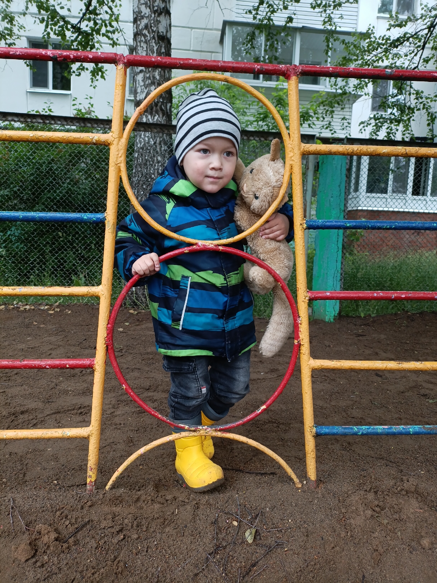 A young child stands within a colorful metal playground structure, holding a plush brown rabbit toy. The child, likely a boy, wears a grey and black striped beanie, a blue and green patterned winter jacket, dark jeans, and bright yellow rain boots. His right hand grips a red bar of the playground equipment, and he looks with a thoughtful or curious expression towards his right. The playground structure itself features red, yellow, and blue painted metal bars, including circular elements, showing signs of wear with chipped paint.
The ground around the structure is dark, damp earth or mud, interspersed with small twigs and debris. In the background, behind a wire fence, are residential buildings, likely apartment blocks, with multiple windows. Green foliage and trees are visible, partially obscuring the buildings and adding to the outdoor setting. The lighting suggests an overcast day, without harsh shadows. The child's attire, particularly the jacket and rain boots, indicates cooler weather, possibly autumn or spring. This scene takes place in a playground setting in Chaikovsky, Russia. No visible text is present in the image.