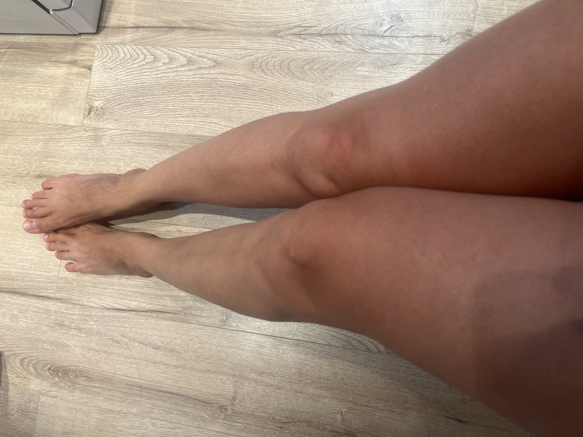 A close-up, overhead shot focuses on a pair of tanned legs and bare feet resting on a light wood-patterned floor. The legs are positioned in a V-shape, with the knees slightly bent and visible. The feet are pointed away from the camera, displaying toes and toenails. The skin on the legs appears smooth with some subtle variations in tone. The flooring is consistent throughout the frame, showing a repeating wood grain pattern.