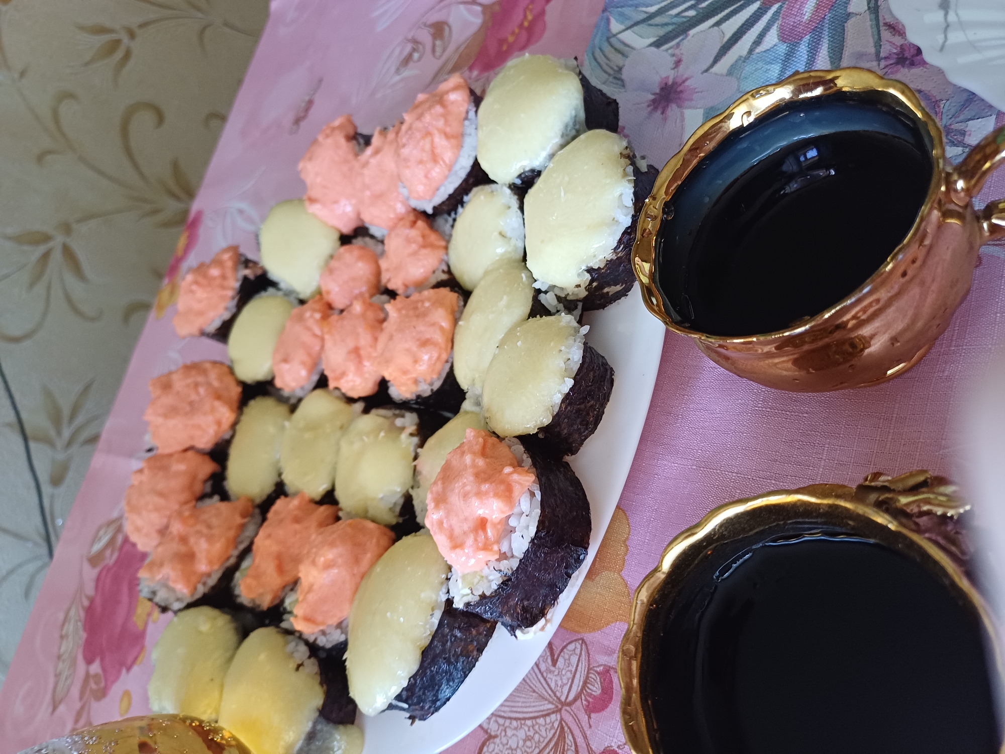A plate of sushi and two cups of dark liquid are arranged on a pink tablecloth with a floral pattern. The sushi appears to be nigiri or maki rolls topped with either a creamy orange sauce or a light yellow topping. The cups are made of a metallic material with a gold rim and have intricate designs on their exterior. The background shows a wall with a patterned wallpaper and a hint of natural light. The overall scene suggests a casual meal setting, possibly at home.