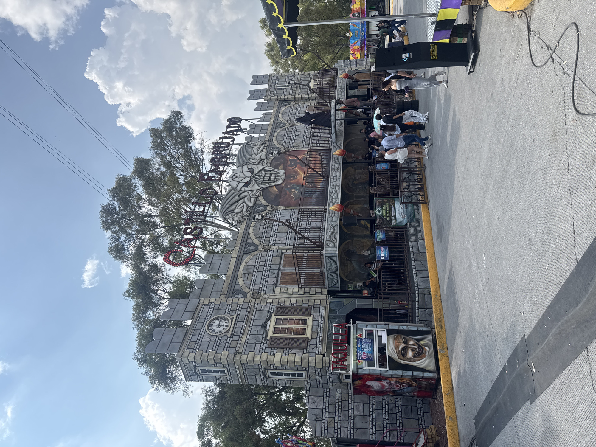 The image depicts a ride named "Mansión del Terror" (Mansion of Terror) at what appears to be a fair or amusement park. The ride is designed to resemble a gray stone castle. People are waiting in line to enter the ride. A black box is to the right of the building. The sky is visible with clouds and trees are around the mansion. The location is likely in Puebla, Mexico.