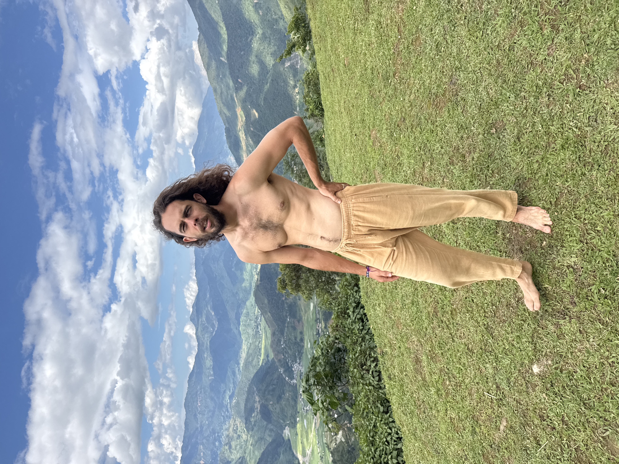 A male, approximately 25-40 years of age, with long, dark, curly hair and a beard, stands centrally on a gently sloping grassy hillside. He is shirtless, revealing his bare chest with visible hair, and wears loose-fitting, tan-colored drawstring pants. A small, dark-colored bracelet is present on his left wrist, and he is barefoot. His left hand is placed on his hip, and his gaze is directed forward towards the camera, with his mouth slightly open. The background presents a panoramic view of a vast, verdant mountainous landscape, overlooking a valley with discernible small settlements and cultivated areas, consistent with the mountainous region of Tú Lệ, Vietnam. The sky above is blue, populated with numerous scattered white cumulus clouds. The scene is illuminated by natural daylight.