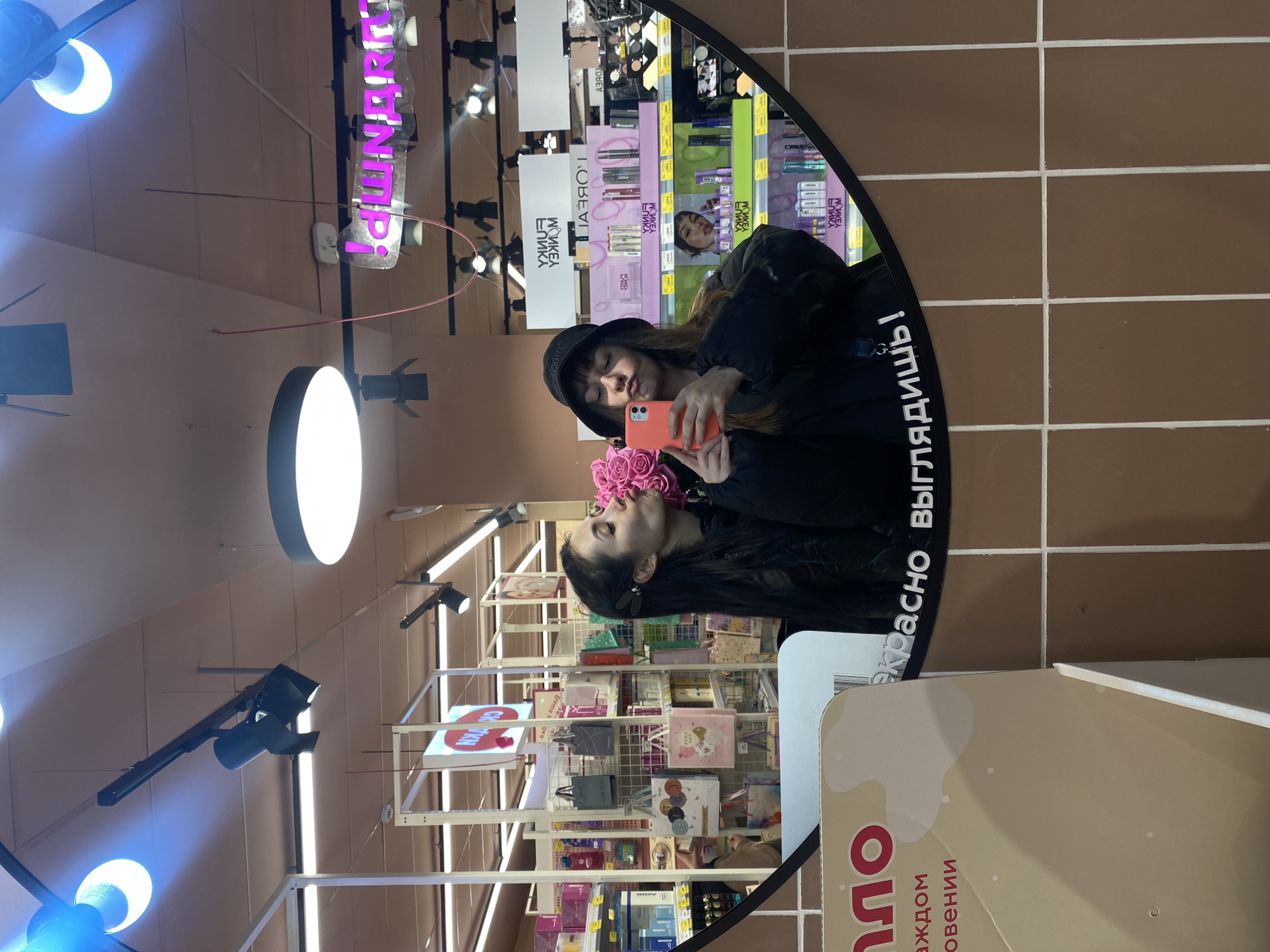 Here is a detailed description of the image:
**Overall Scene:**
The image shows a reflection in a mirror within a retail setting, possibly a store. The mirror is curved, adding a distorted perspective. The background indicates a retail environment with shelves stocked with various goods.
**People:**
Two people are discernible:
* A woman is taking a selfie in the mirror. She wears a dark hat and dark clothing and holds a pink mobile phone. Her facial expression seems playful, possibly a 'duck face'.
* Her reflection appears on the other side of the mirror, and she is holding a bouquet of pink flowers.
**Objects:**
* Curved Mirror: This is the primary element framing the shot.
* Pink Bouquet: The woman holds a bouquet of pink flowers, possibly roses.
* Mobile Phone: She's holding a mobile phone, presumably to take the selfie.
* Retail Shelves: Behind the woman in the mirror, there are shelves stocked with products, indicating a retail environment.
* Lighting Fixtures: Overhead lighting fixtures are visible, including bright circular lights and spotlights.
* Product Packaging: Various retail products are visible on the shelves, with brands and packaging distinguishable.
**Actions/Interactions:**
* Selfie-Taking: The primary action is the woman taking a selfie in the mirror.
* Retail Environment: The presence of products on shelves suggests customers browsing or shopping.
**Environment:**
* Retail Store: The presence of stocked shelves and lighting suggests a retail store or department.
* Walls: The wall surrounding the mirror has an earth tone tile and the writing 'красно выглядишь!', which translates to 'look good!'.
**Location:**
The products on the shelves include brands like L'Oreal and Funky Monkey, indicating an availability of cosmetics. The sign 'пло' with the text 'рядом с новении' means 'near novelties'. The name in the mirror reflection 'ЕЛИКА' can be a store name. The wall surrounding the mirror says, 'красно выглядишь!'.
Based on the information available, it is difficult to confirm, but it may be located in Votkinsk, Russia.
