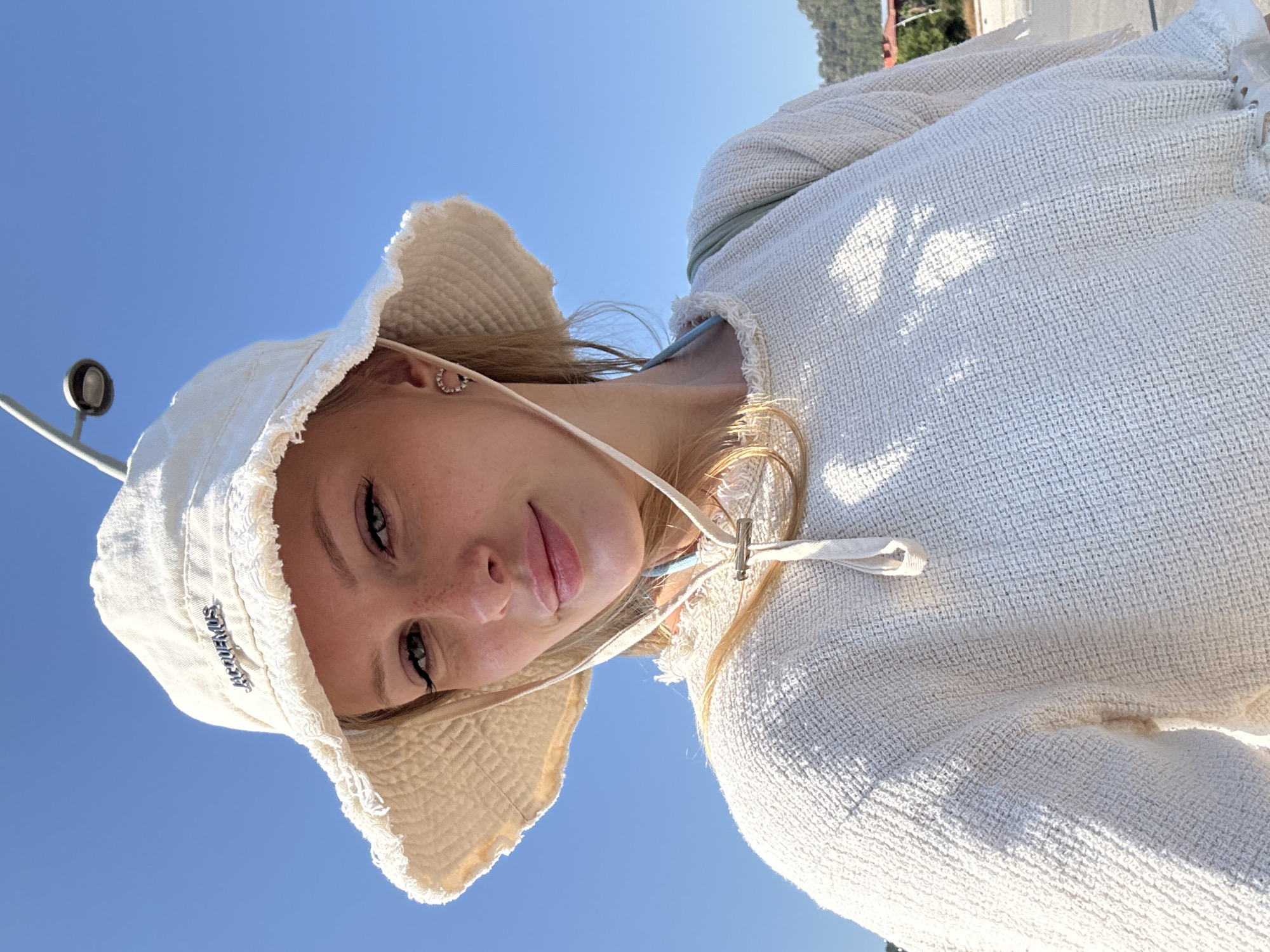 A young woman, wearing a white bucket hat and a white textured sweater, smiles at the camera. She has blonde hair and is wearing a small, circular earring. The hat has a frayed brim and a silver logo that appears to read "JACQUEMUS". A light blue strap is visible under her sweater. The background is a clear, bright blue sky. A street lamp is visible in the upper right corner of the image. The scene appears to be outdoors, possibly in a sunny, coastal location, given the attire and clear sky. The background also shows hints of distant trees and buildings, suggesting an urban or semi-urban environment. No specific landmarks in Kemer, Turkey are identifiable from the image.