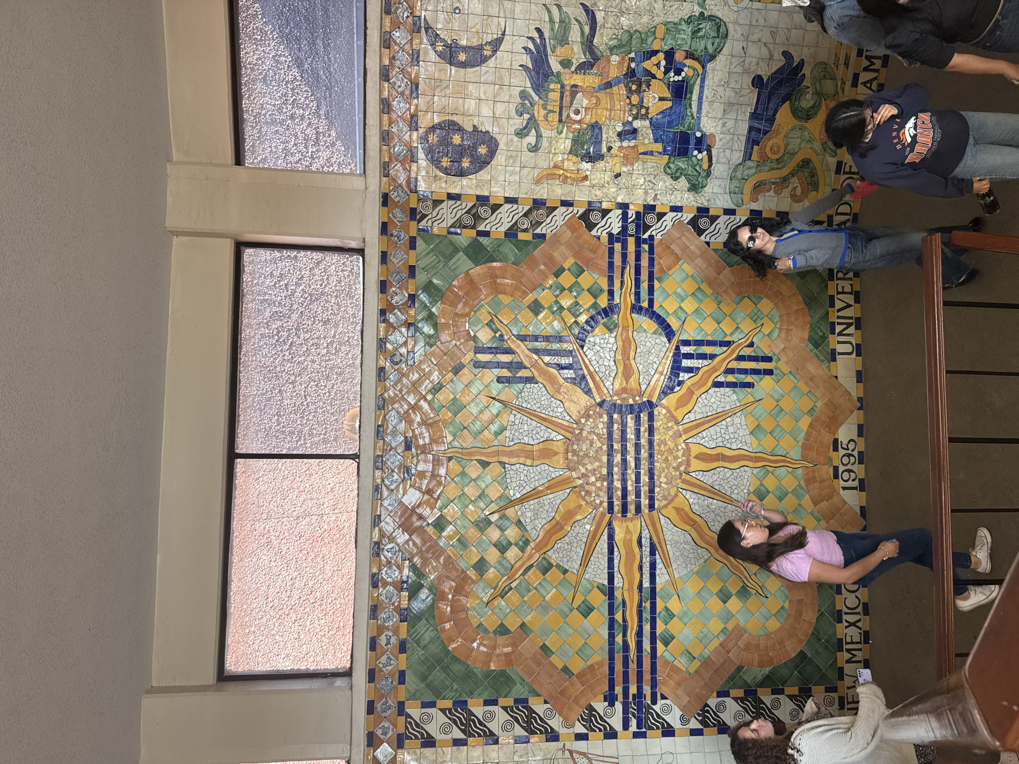 The scene depicts an interior space in San Andrés Cholula, Mexico, featuring a group of five individuals on a descending staircase or ramp adjacent to a large, colorful tiled wall mosaic.
The mosaic dominates the wall, displaying a central sun-like emblem with rays in warm tones, set against a background of green and yellow checkerboard patterns, interspersed with geometric and wave-like motifs. To the right of the sun, a multi-colored depiction of an anthropomorphic figure, possibly a Mesoamerican deity, is visible, with two crescent moons and stars above it. Along the lower edge of the mosaic, partial inscriptions read "NEW MEXICO", "1995", and "UNIVER". Above the mosaic, multiple rectangular windows with dark frames contain textured or frosted glass, allowing diffused light into the space. A plain, light-colored, textured ceiling is visible above the windows. The floor is dark, possibly concrete, and a brown metal handrail marks the side of the descent.
Five individuals, primarily young women, are observed.
1. On the far left, a female with curly brown hair wears a light-colored cardigan over a patterned top. She holds a white smartphone in her left hand and looks to her left.
2. Next to her, a female with straight dark hair and glasses wears a light purple short-sleeved top, dark blue jeans, and white sneakers. She is depicted bringing a clear bottle or cup to her mouth, looking to her right.
3. In the center, a female with long dark wavy hair wears dark sunglasses, a grey long-sleeved cardigan with blue trim over a dark top, and light blue jeans with dark shoes. She looks towards the viewer, holding a red object, possibly a bag or scarf, in her left hand.
4. To her right, a female with dark hair wears a navy blue sweatshirt bearing "DENVER BRONCOS" text and emblem, and light blue jeans. She holds a dark bottle in her right hand and looks downwards.
5. On the far right, a fifth individual is partially visible, wearing dark clothing and dark blue jeans, also descending the stairs.
All individuals are in various stages of walking downwards on the staircase or ramp.