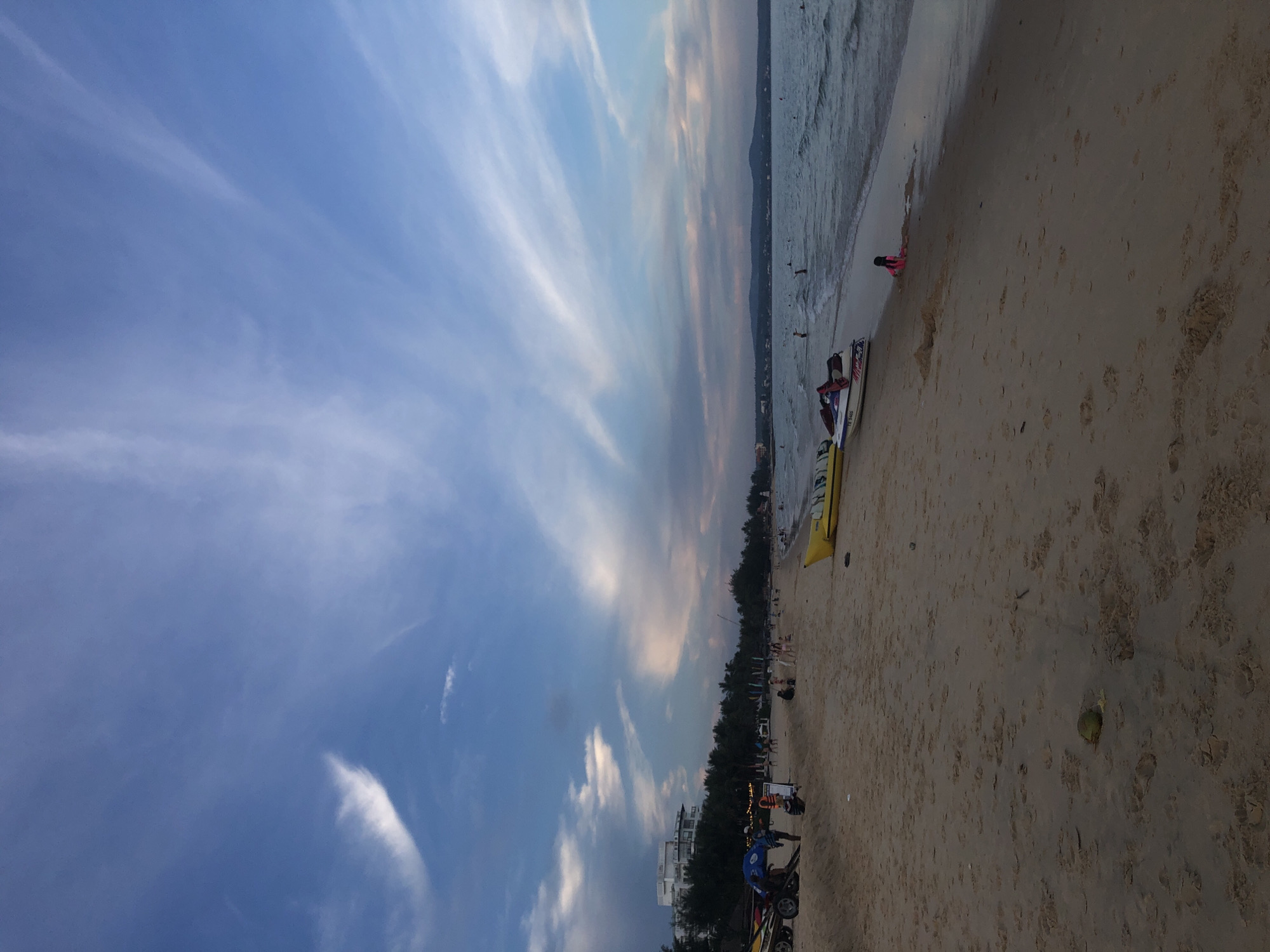 The image depicts a beach scene in Mũi Né, Vietnam, during what appears to be late afternoon or early evening, given the soft lighting and the sky. The foreground is dominated by a sandy beach with tire tracks and footprints, indicating recent activity. A green coconut lies on the sand towards the bottom left.
Moving towards the middle ground, several people are scattered along the beach. Some are walking, while others are seated or standing. A blue tarp is set up on the left, with people gathered around it. A yellow banana boat is positioned on the sand, and a jet ski is nearby. Further down the beach, a child in a pink swimsuit is playing near the water's edge.
The middle distance shows the ocean with gentle waves and several people swimming or wading in the water. A shoreline is visible in the far distance, featuring a line of palm trees and buildings, suggesting the presence of a resort or town. The sky is a prominent feature, with a mix of blue and wispy white clouds, some tinged with pink and orange, indicative of sunset. The overall atmosphere is relaxed and recreational, typical of a beach resort area.