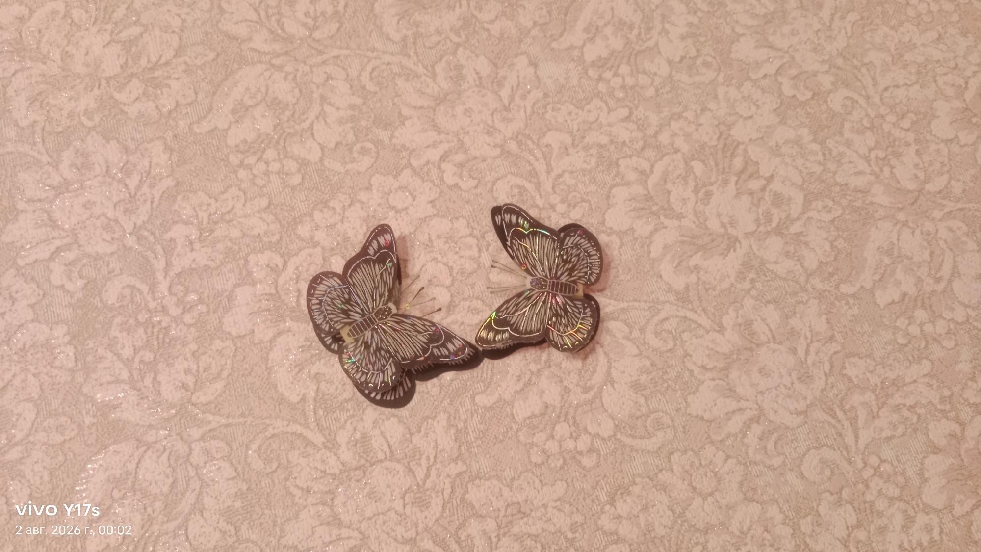 The image shows two decorative butterflies with iridescent wings against a pale pink, floral-patterned textured background. The butterflies are positioned centrally, with one slightly to the left and the other to the right, both facing towards the viewer's right. The background is a wallpaper with a raised floral design, possibly a damask pattern, and appears to have a subtle shimmer. The bottom left corner of the image contains a watermark indicating "vivo Y17s" and the date "2 авг. 2026 г., 00:02", which translates to August 2, 2026, 00:02. There is no visible indication of the city of Rudny, Kazakhstan, in the image itself.