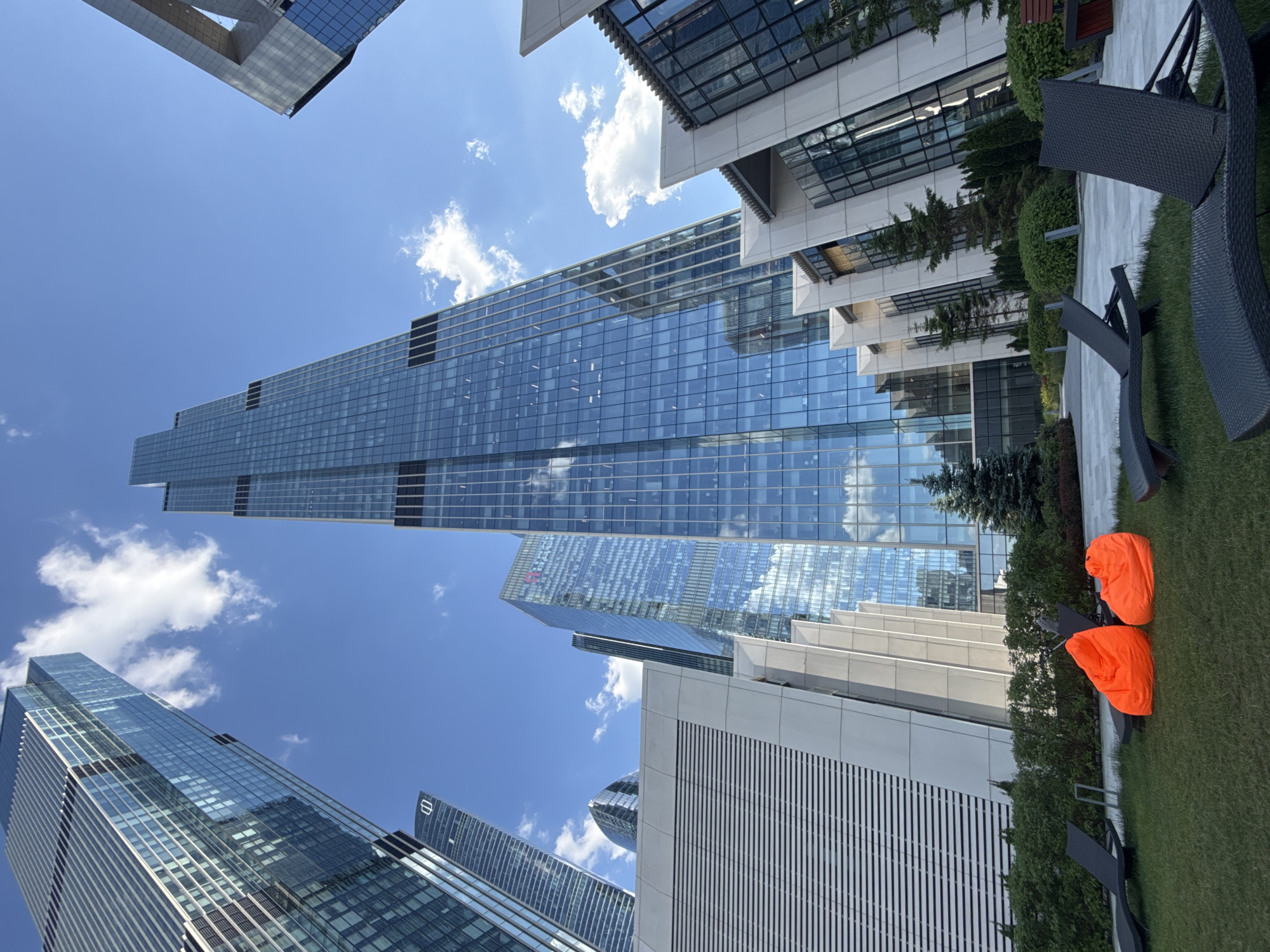 This image captures a modern urban landscape in Moscow, Russia, under a clear, sunny blue sky with scattered white cumulus clouds.
The scene is dominated by several contemporary glass skyscrapers that reflect the expansive blue sky and other surrounding buildings. A particularly tall and slender glass skyscraper stands prominently in the center of the frame, its reflective facade showing a mosaic of blues and whites. Other angular, multi-story glass structures are visible around it, contributing to a sense of a bustling business district.
On the right side of the image, there is a landscaped area designed for relaxation. It features vibrant green grass, neatly trimmed hedges, and small evergreen trees, suggesting a well-maintained park, rooftop garden, or plaza. Dark-colored, wicker-style outdoor lounge chairs are arranged on the grass. Two of these chairs are notably covered with bright, almost fluorescent orange, sack-like objects, which could be protective covers for cushions or decorative bean bags. Light-colored paved sections are also visible within this green space.
The time of day appears to be bright daylight, likely midday or early afternoon, given the clear sky and strong sunlight. There are no people visible in the frame. A notable detail is a stylized "8" logo visible on the upper section of one of the glass buildings on the far left, providing a potential location cue. The overall impression is one of modern architectural design harmoniously integrated with accessible green spaces in a metropolitan setting.