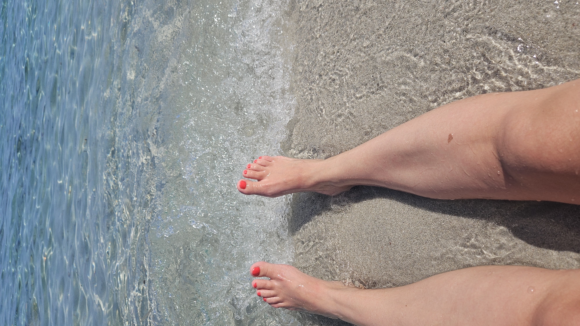 This is a low-angle, full-frame shot of a person's bare legs and feet at the edge of the sea in L'Île-Rousse, France. The feet are positioned on wet sand, with the toes pointing upwards. The toenails are painted a vibrant coral color. Small waves are lapping at the feet, with clear, shallow water covering the sand. The background consists of the sparkling blue sea, with sunlight glinting off the surface. The sand is light-colored and appears fine-grained. The overall impression is one of relaxation and enjoyment of a sunny day at the beach.