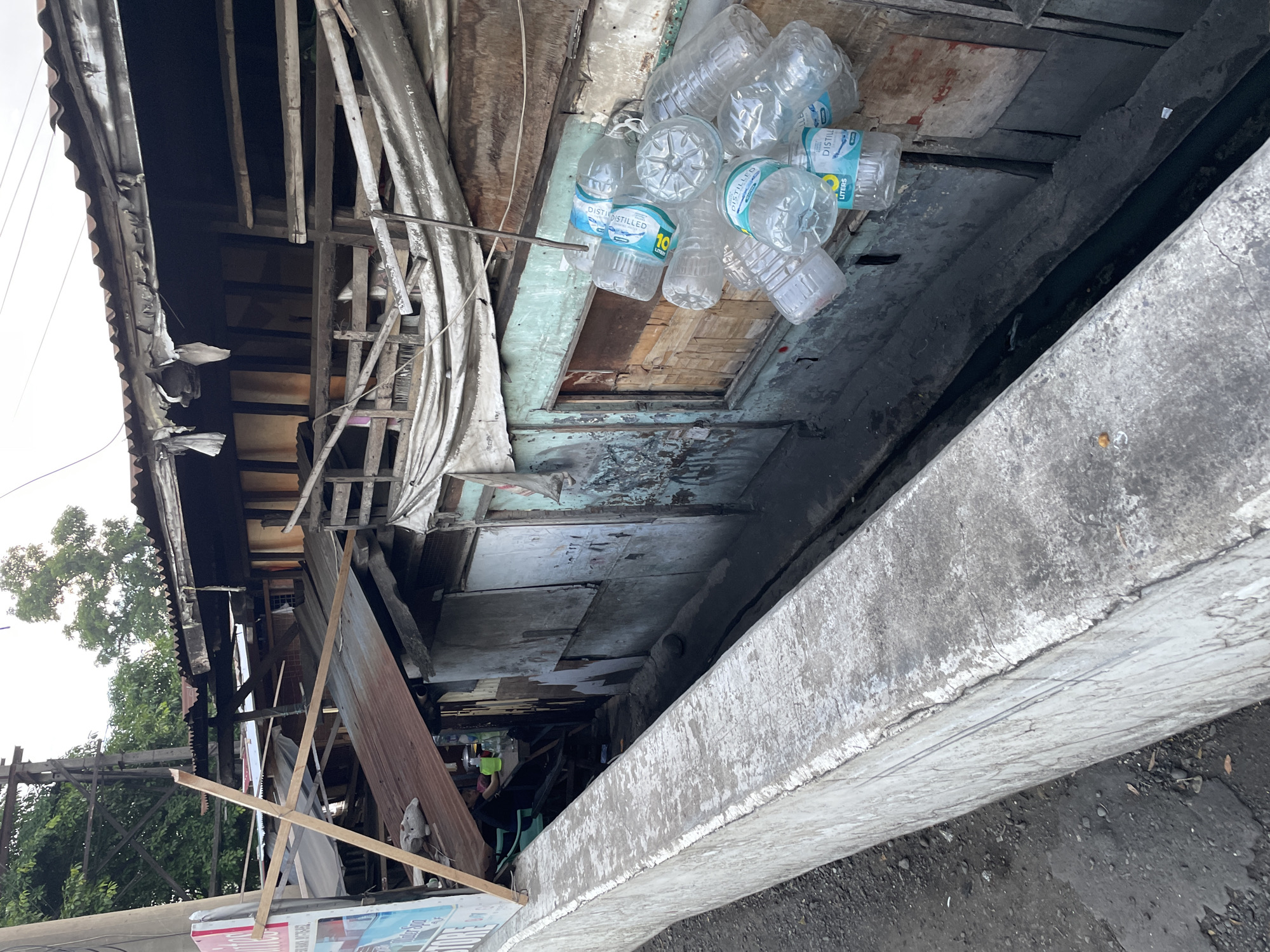The image shows a dense urban environment, possibly a slum area in Davao City, Philippines, characterized by ramshackle structures constructed from corrugated metal, wood, and tarps. The buildings appear to be multi-story, with precarious wooden balconies and supports. A concrete barrier runs along the foreground, separating a dark, narrow waterway from what appears to be a street or pathway.
A prominent cluster of empty plastic water bottles hangs from a wire, suspended against one of the weathered wooden walls of a structure. The bottles are mostly clear, with some exhibiting blue and white labels indicating "Distilled" and "Liters." The surrounding buildings display signs of disrepair, with peeling paint, exposed wood, and patched roofing. In the background, trees and utility poles are visible, suggesting the presence of infrastructure within the densely packed settlement. The overall scene depicts a scene of poverty and resourcefulness, with discarded items being repurposed or collected.
